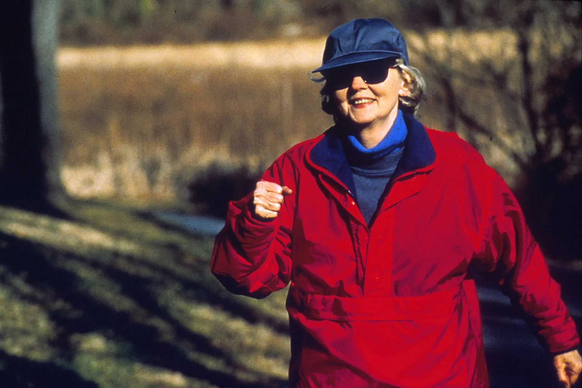 A smiling older woman in a red jacket, cap and sunglasses power walking along a sunlit path.