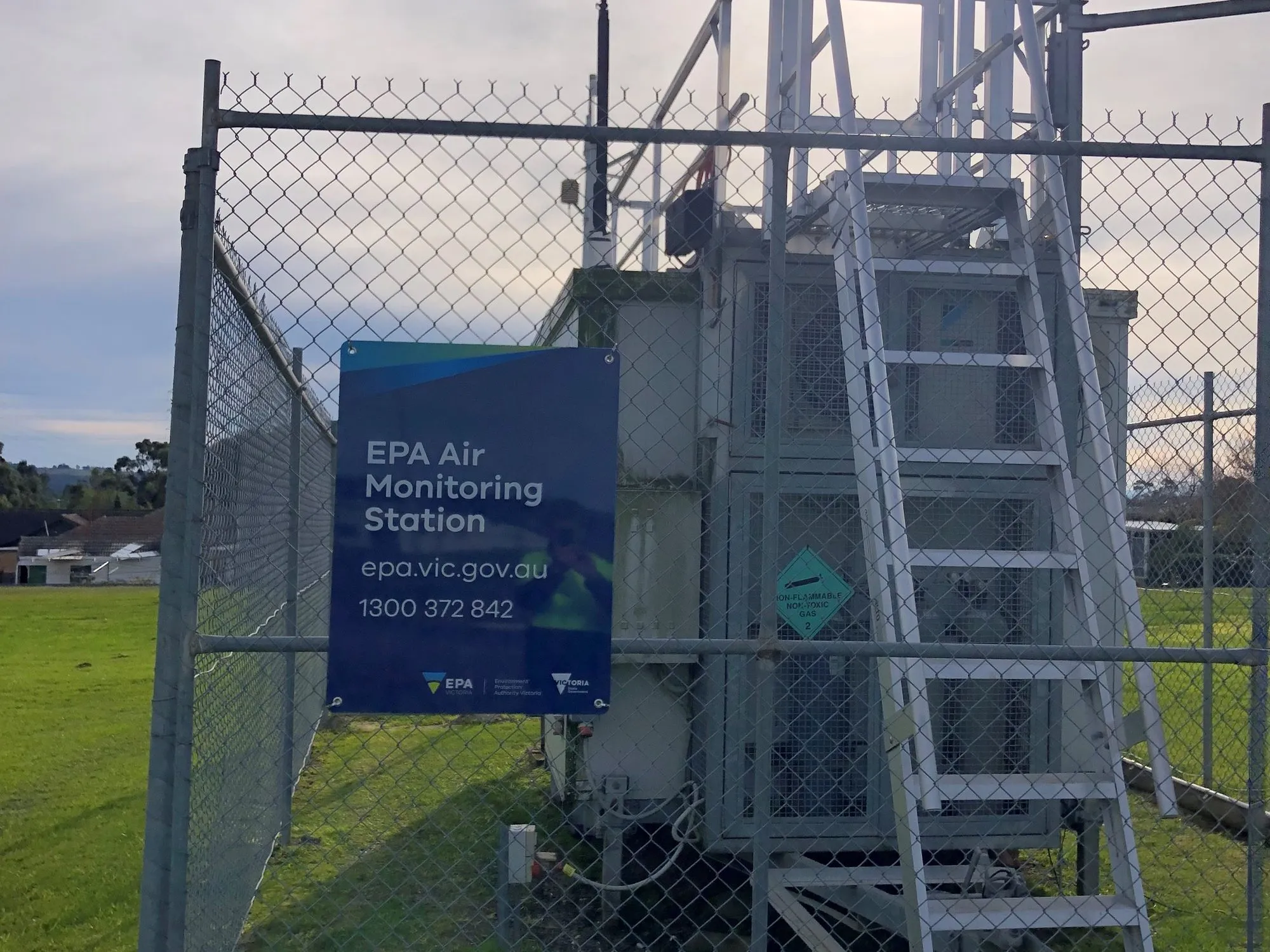A fenced air monitoring station with instruments and a sign reading EPA Air Monitoring Station on a grassy site.