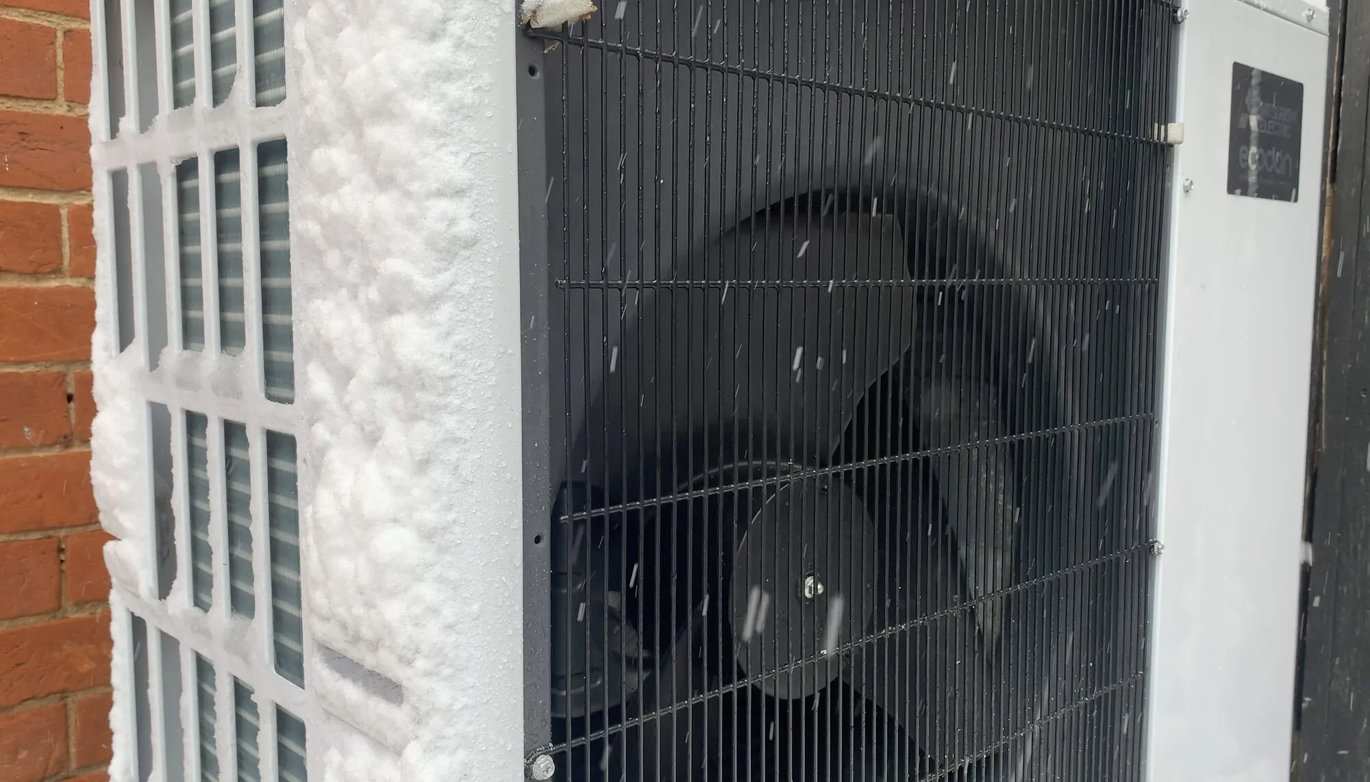 The outdoor unit of an air-source heat pump, with a large fan behind a grille, its casing and the wall beside it covered in snow.