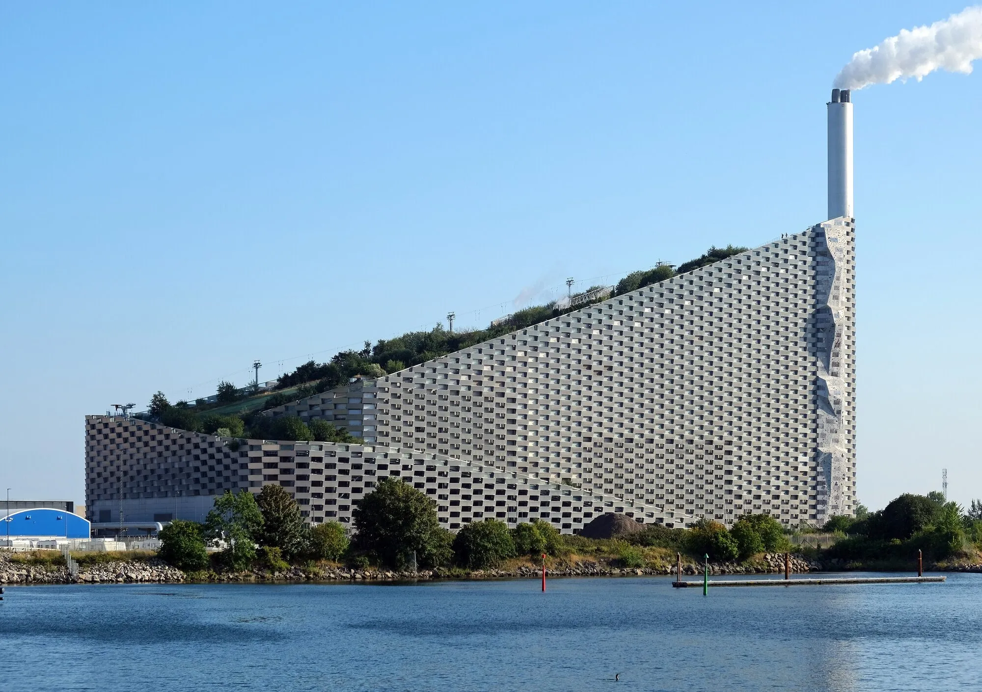 A long, sloping power plant clad in aluminium, with a green ski slope on its roof and a tall chimney, beside the water in Copenhagen.