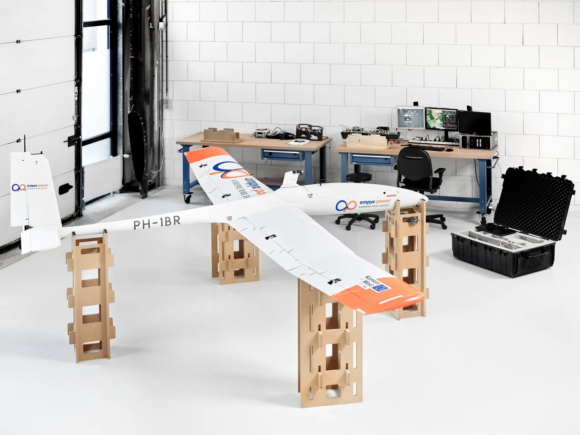 A white rigid glider with an orange and blue stripe resting on cardboard stands in a bright workshop, with computers and equipment cases behind it.