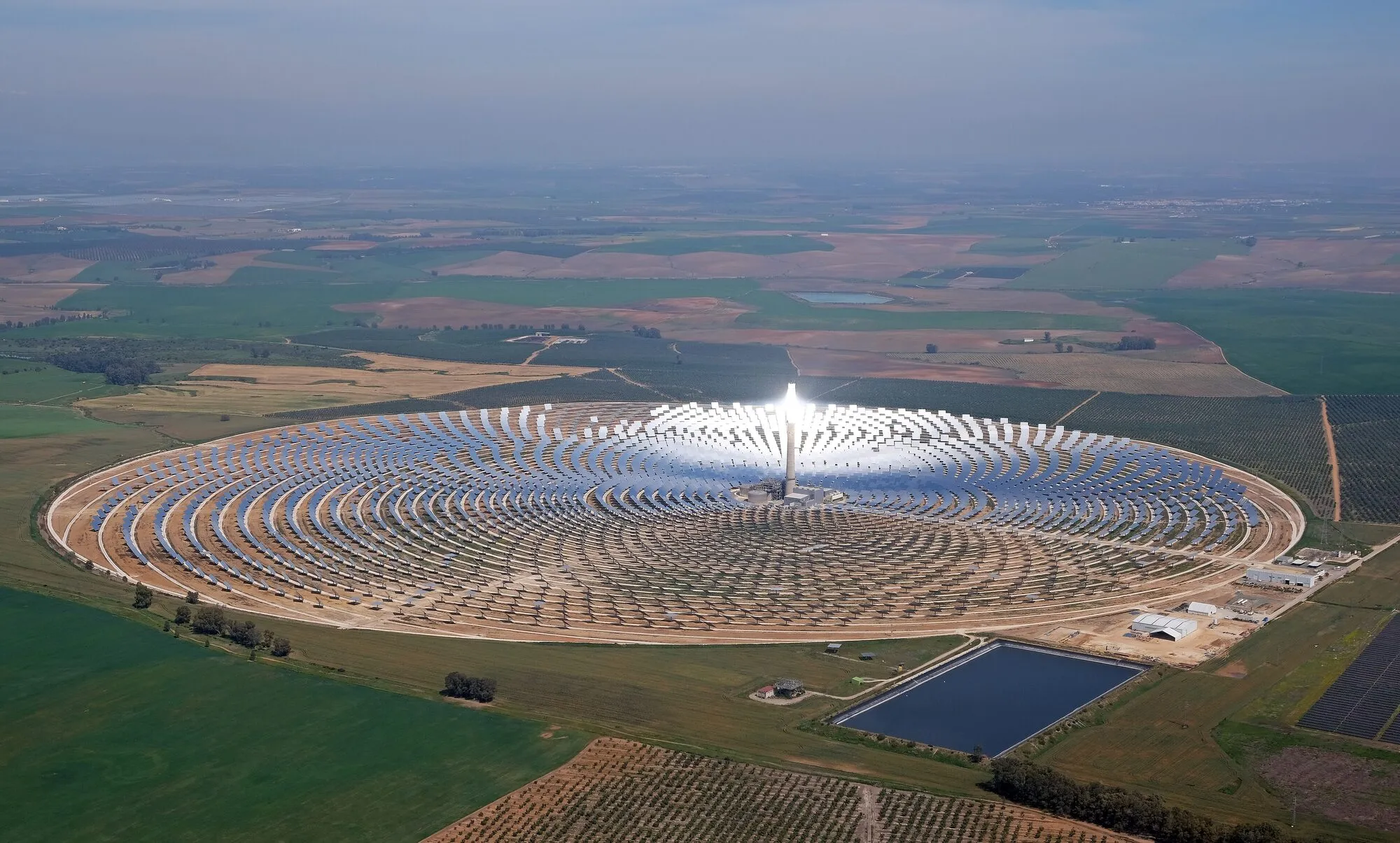 A vast circle of mirrors in green Andalusian farmland, all inclined towards a central tower whose top shines white with reflected sunlight.