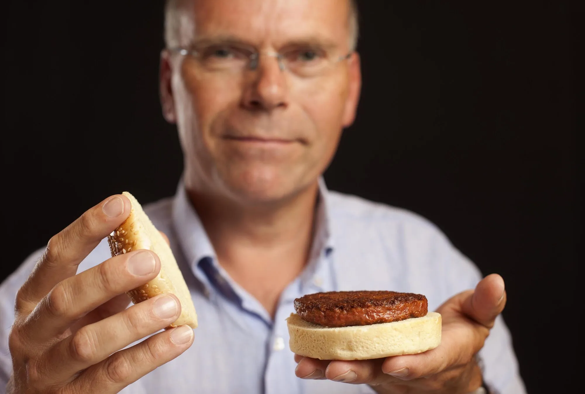 Un home amb una camisa blava sosté un panet amb una hamburguesa cuita en una mà i la part superior del panet a l'altra.