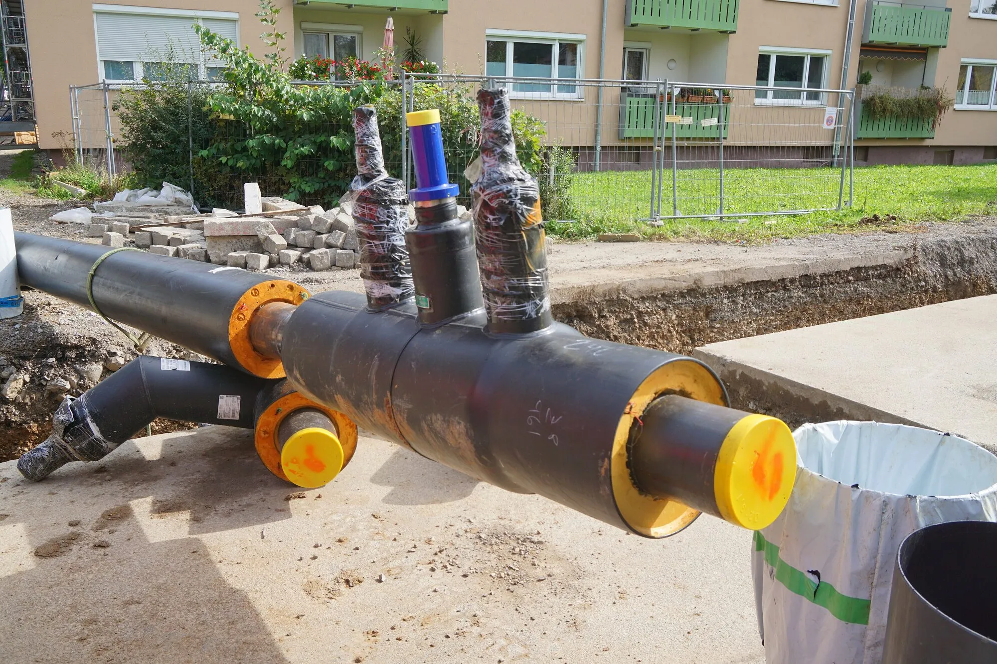 A section of black insulated district heating pipe with branch connections and yellow end caps, beside a trench in front of a block of flats.