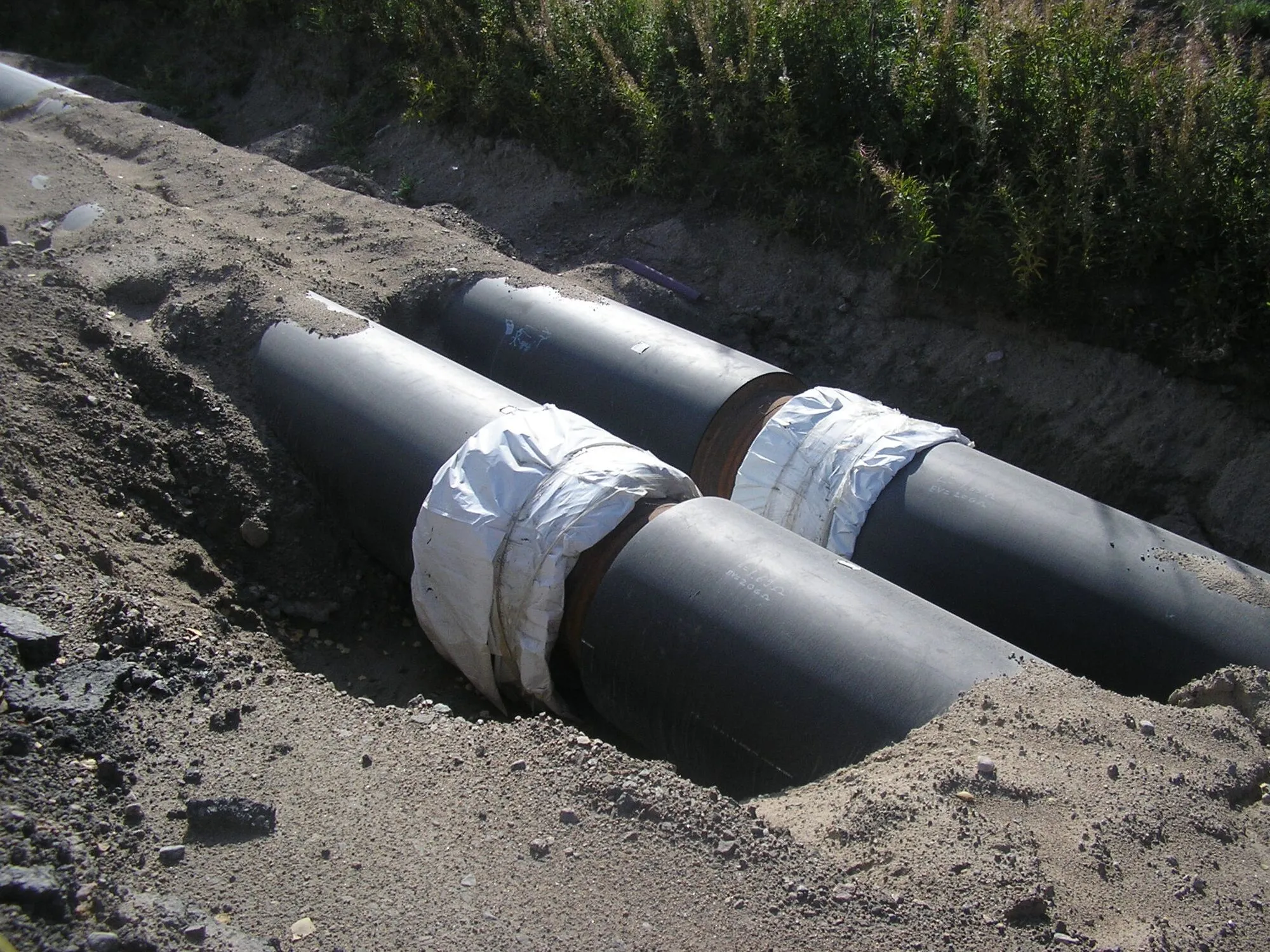 Two large black insulated district heating pipes lying in a freshly dug trench.