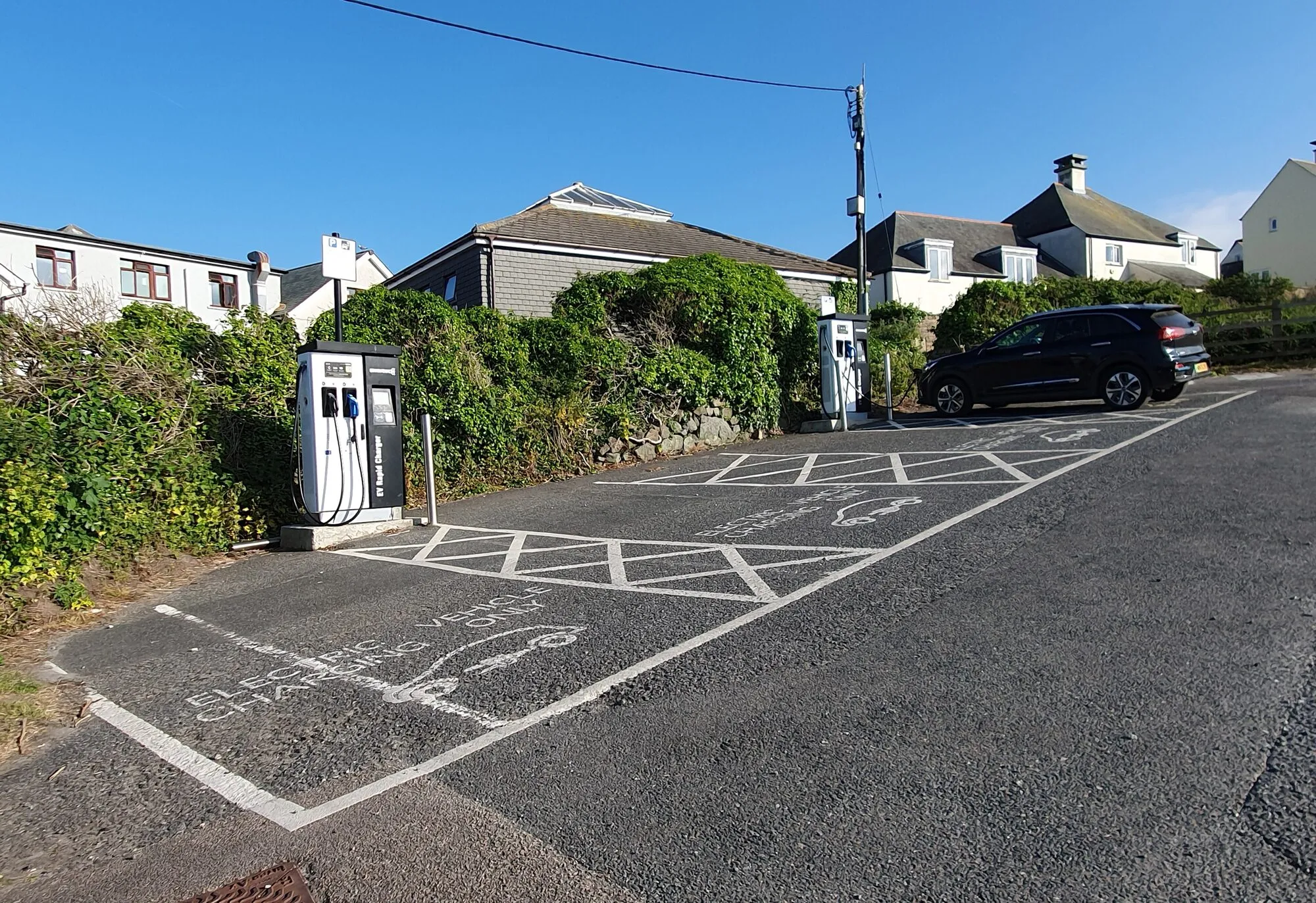 Two electric car charging posts beside marked parking bays in a car park, with houses behind.