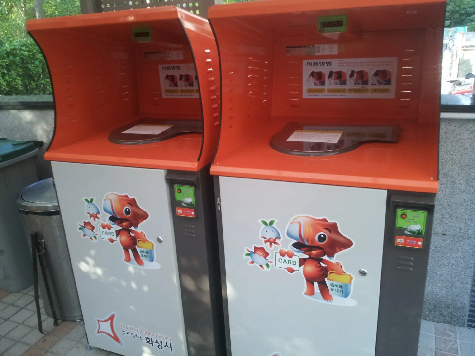 Dues papereres taronges i blanques de residus alimentaris en un carrer, cadascuna amb un lector de targetes, una pantalla i mascotes de dibuixos animats al davant.
