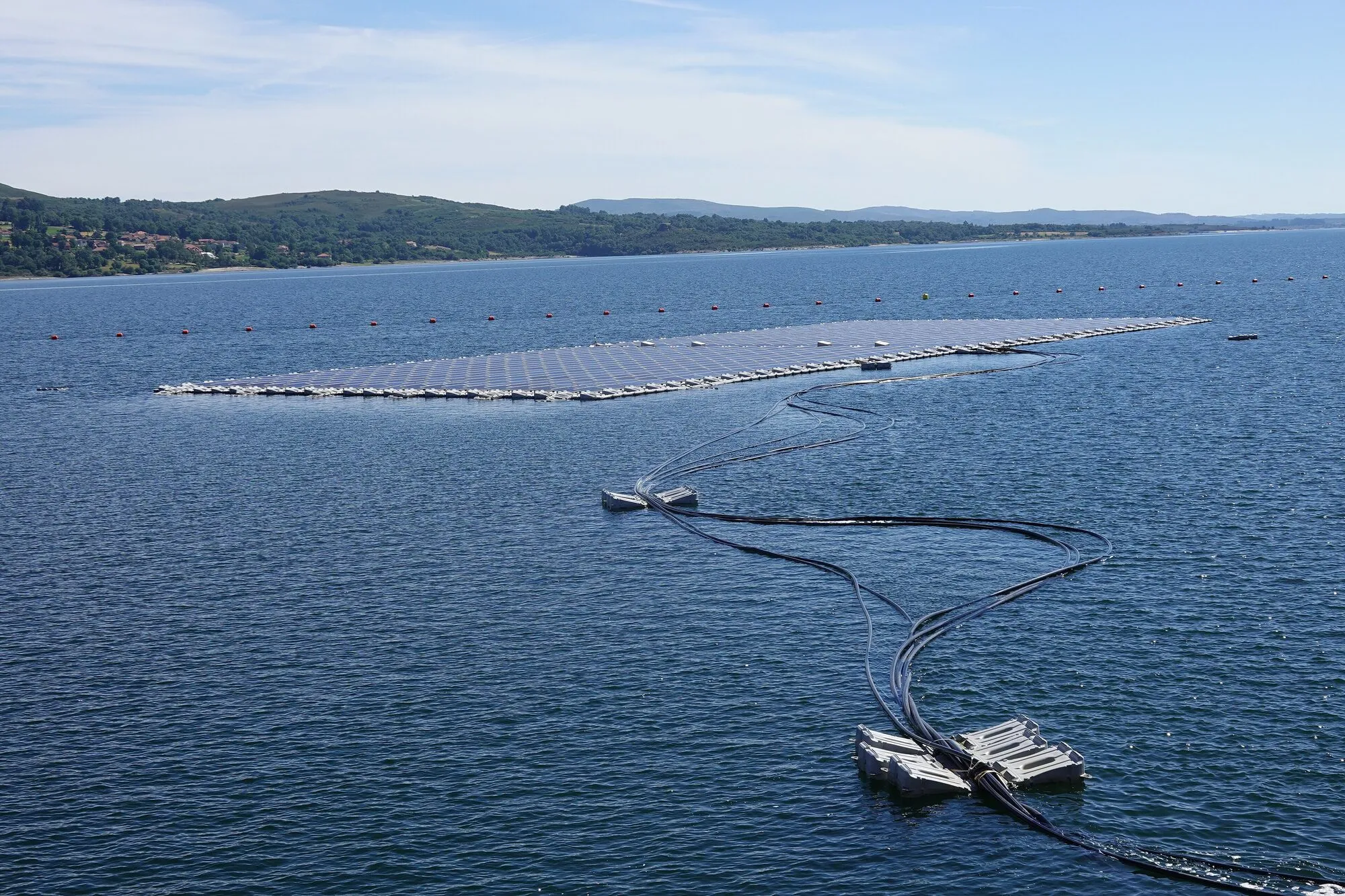 A rectangular floating solar array on a wide blue reservoir, with bundles of black power cables snaking across the water on small floats towards the camera.