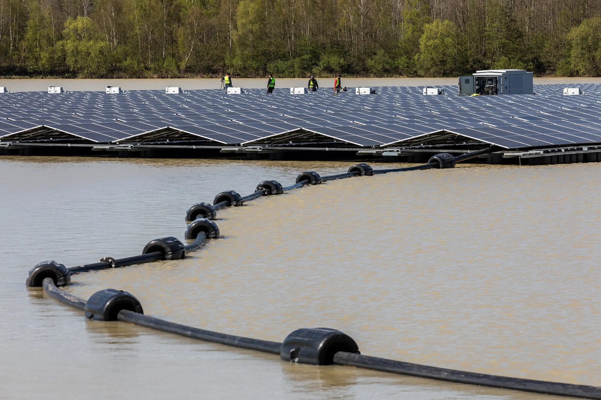 Black floating booms curve across a sandy quarry lake in front of a large array of solar panels, with workers on the array and woodland behind.