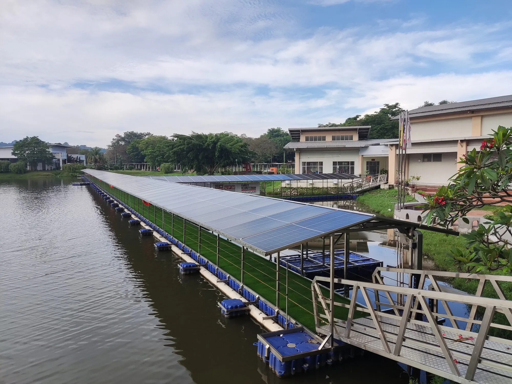 Rows of solar panels on white plastic floats beside a covered walkway that runs out from a lakeside building.