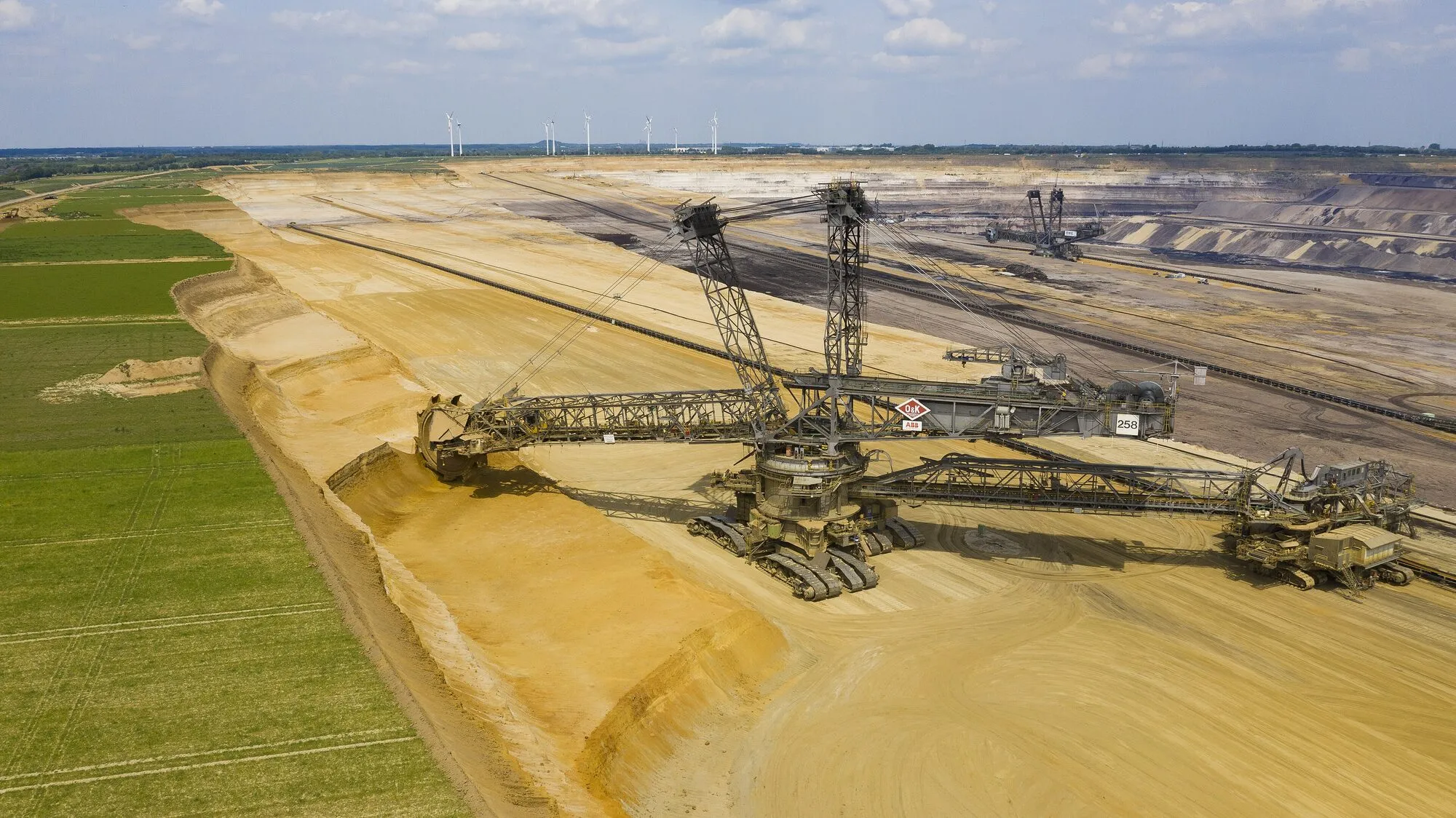 Two giant bucket-wheel excavators in a vast sandy open-cast mine, with green fields and a row of wind turbines on the horizon.