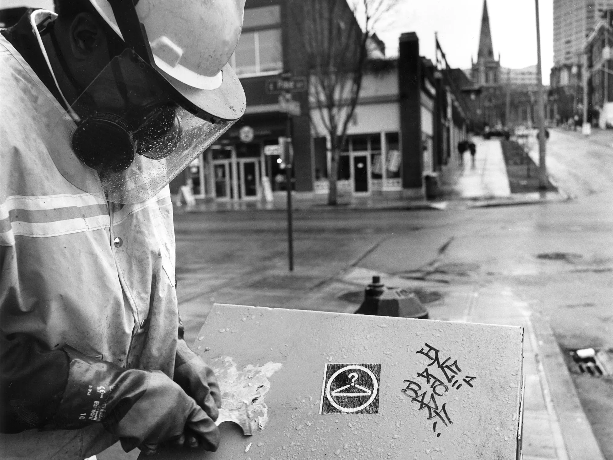 'n Stadswerker met 'n harde hoed en respirator wat 'n graffiti-etiket van 'n nutsdoos op 'n straathoek afskrop, in swart en wit.