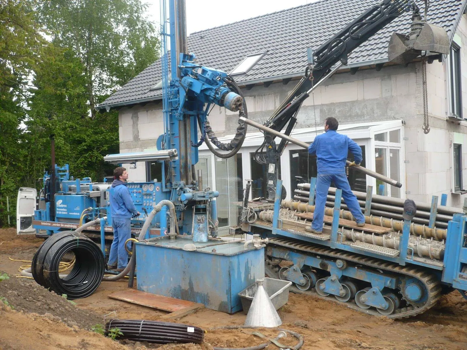 A tracked drilling rig with a tall mast drilling a borehole beside a house under renovation, with two workers and a coil of black plastic pipe.
