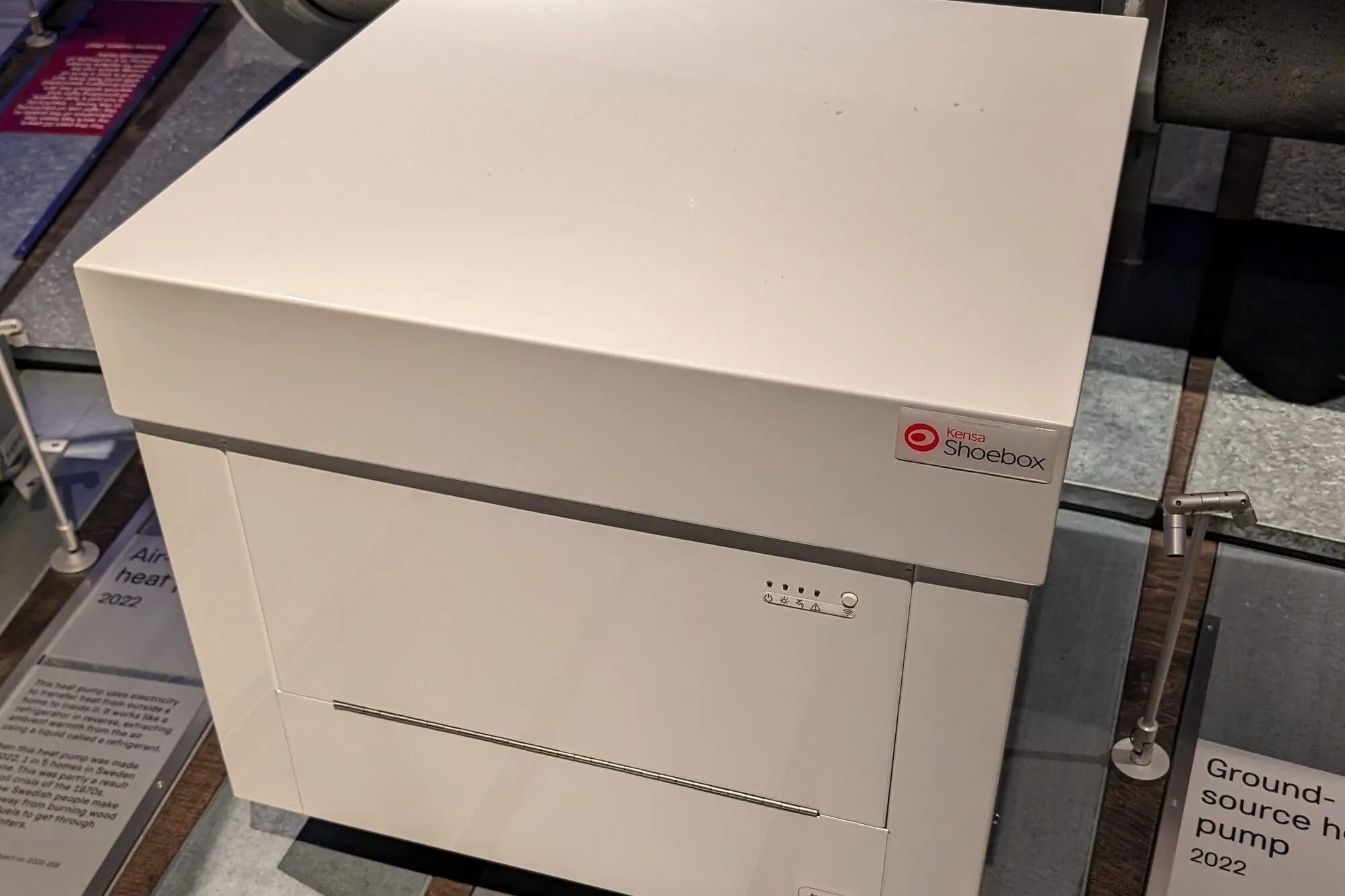 A compact white ground-source heat pump, the size of a small cabinet, on display in a museum.