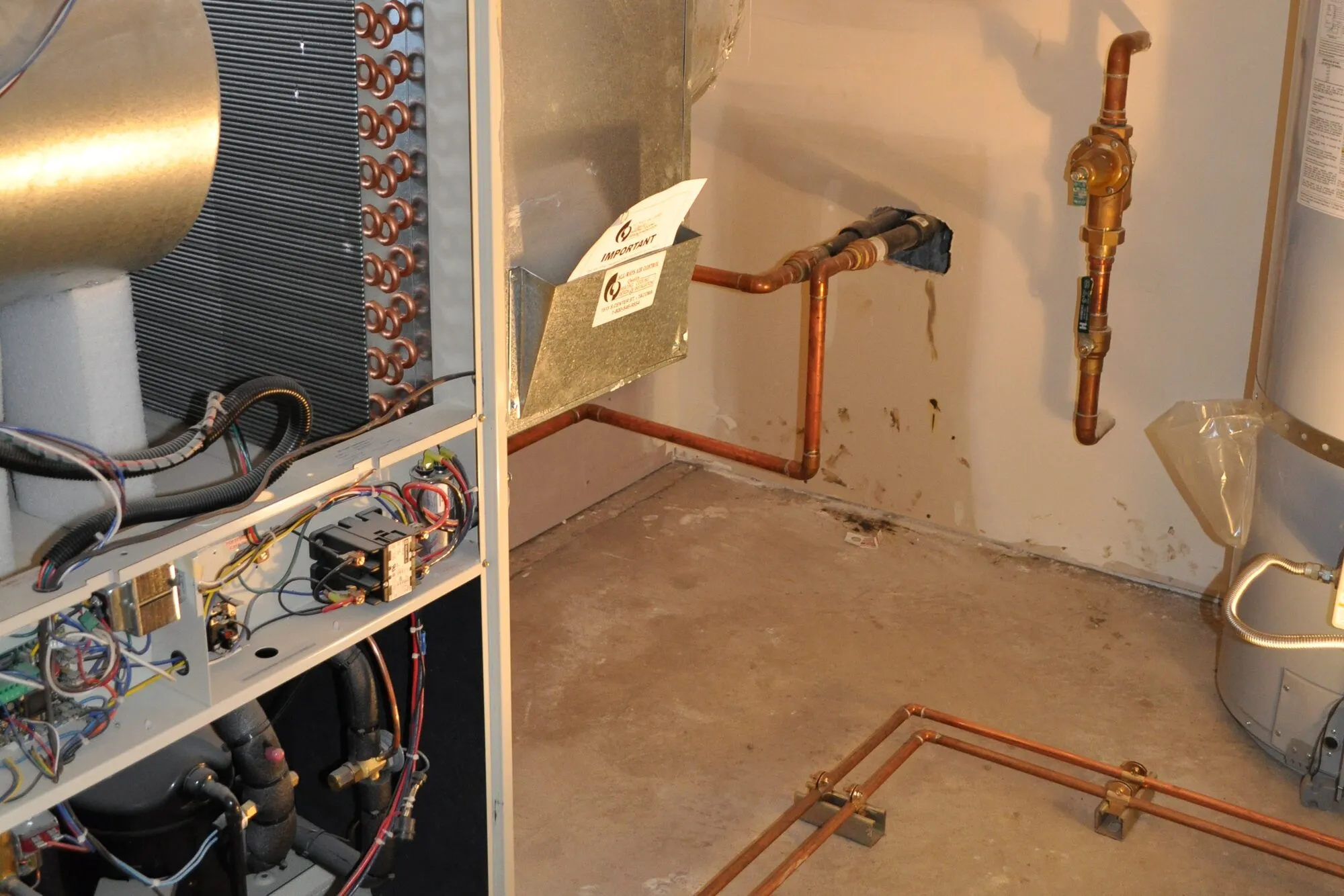 The open cabinet of a home heat pump, with its copper coil and wiring, beside copper pipework on a basement wall.