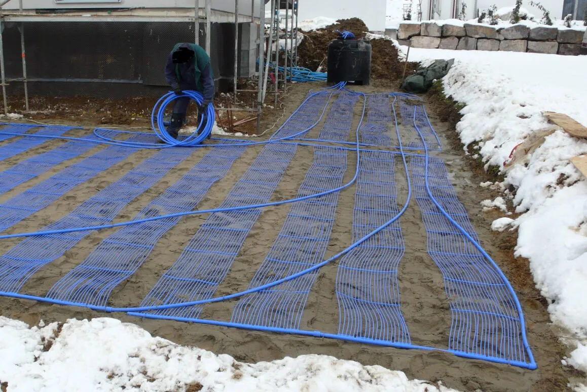 Workers laying rows of blue plastic pipe across a wide excavated area next to a building, with snow at the edges.