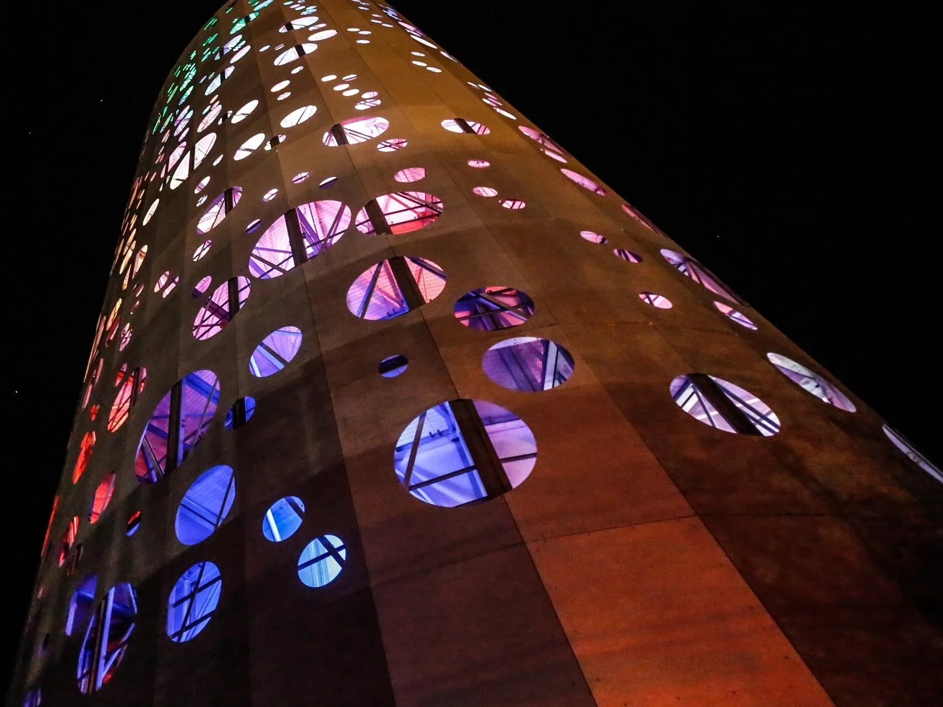 Looking up at a tall cylindrical tower at night, its rust-coloured cladding pierced by round windows glowing blue, pink and white.