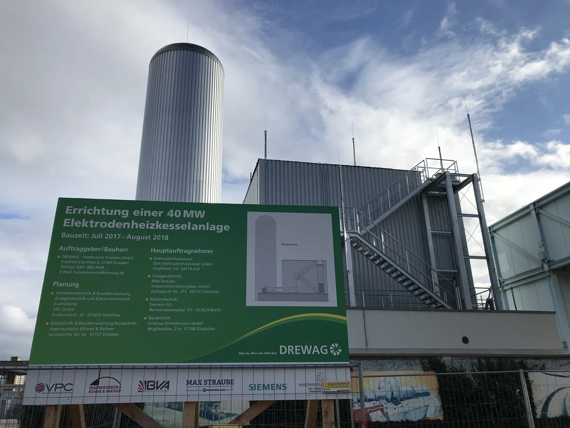 A construction site with a tall silver cylindrical tank and a new plant building behind a large green sign announcing a 40 MW electrode boiler plant.