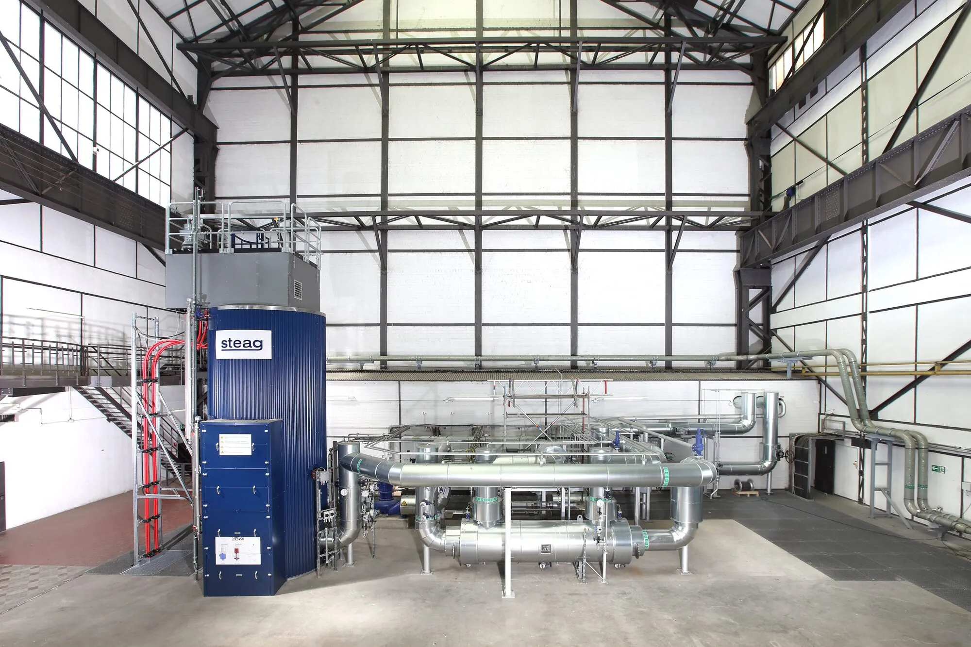 Inside a tall industrial hall: a blue cylindrical boiler with insulated pipework, valves and walkways.
