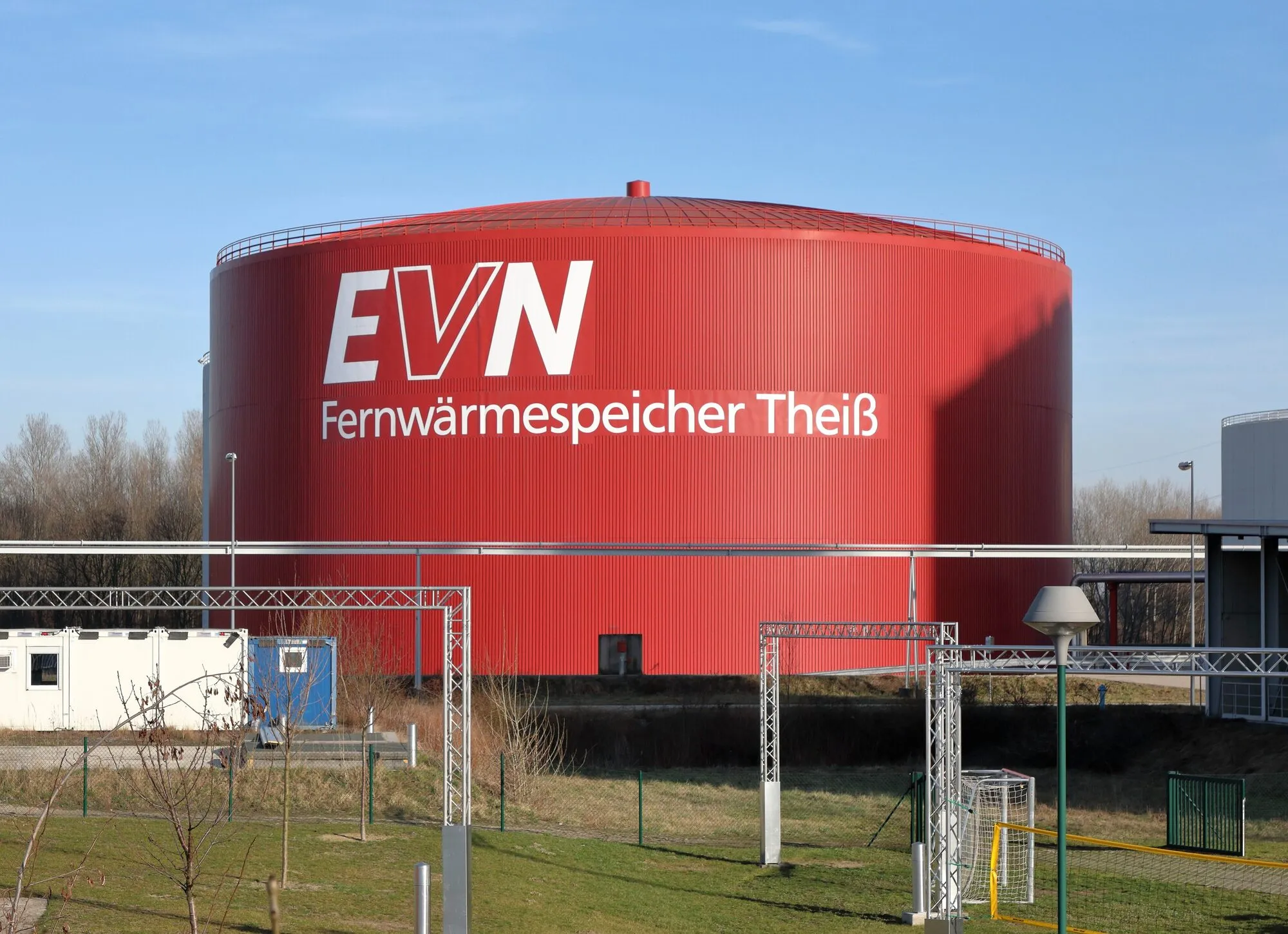 A large red cylindrical tank labelled Fernwärmespeicher Theiß, surrounded by pipe racks.