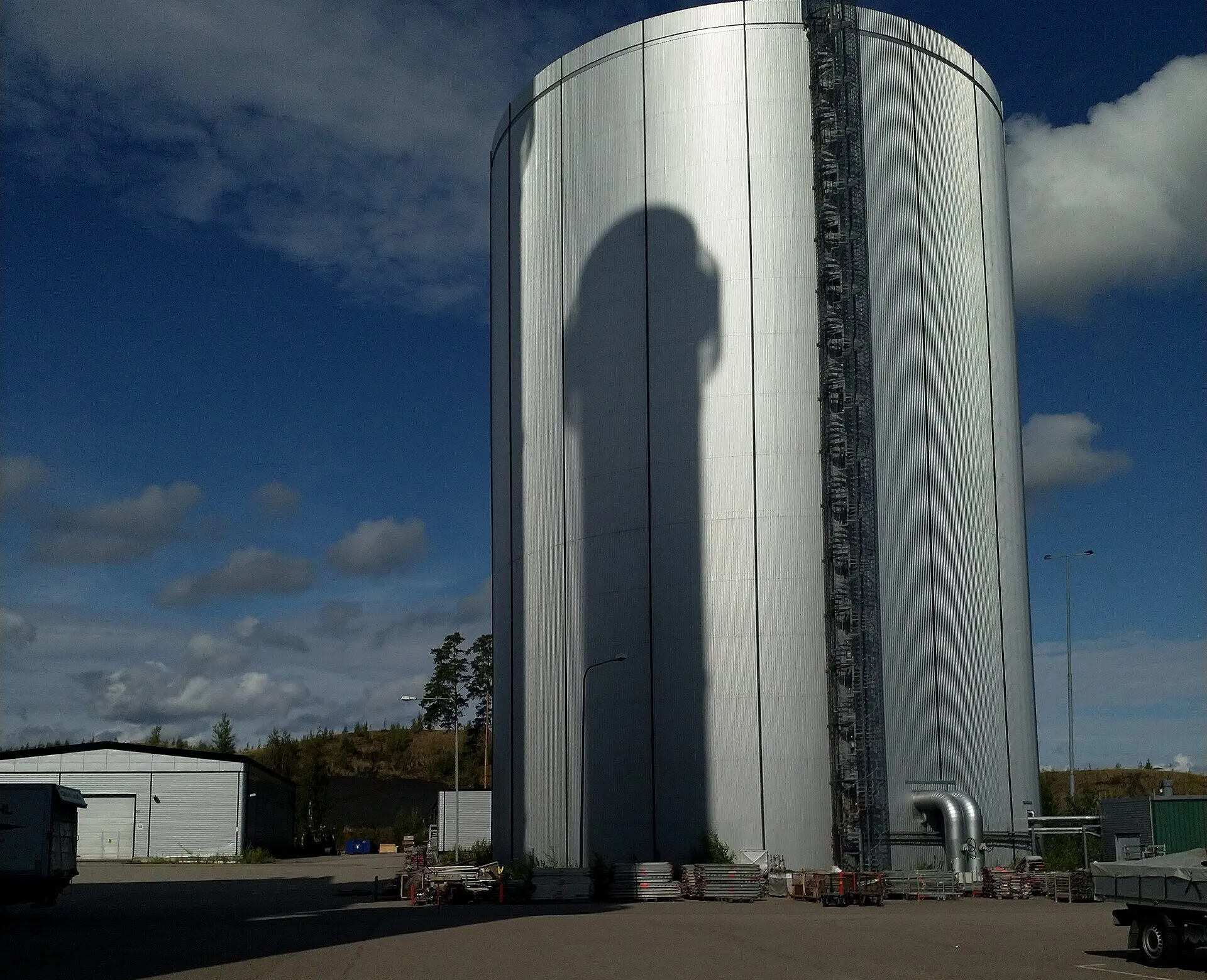 A tall, round, silver-clad storage tank against a deep blue sky, standing on an empty paved yard.