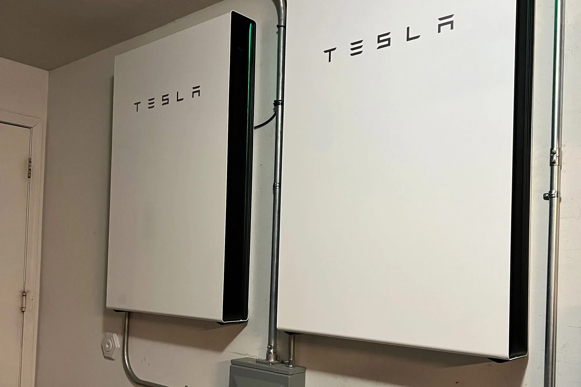 Two white home batteries mounted on a garage wall, with a small switch box below.