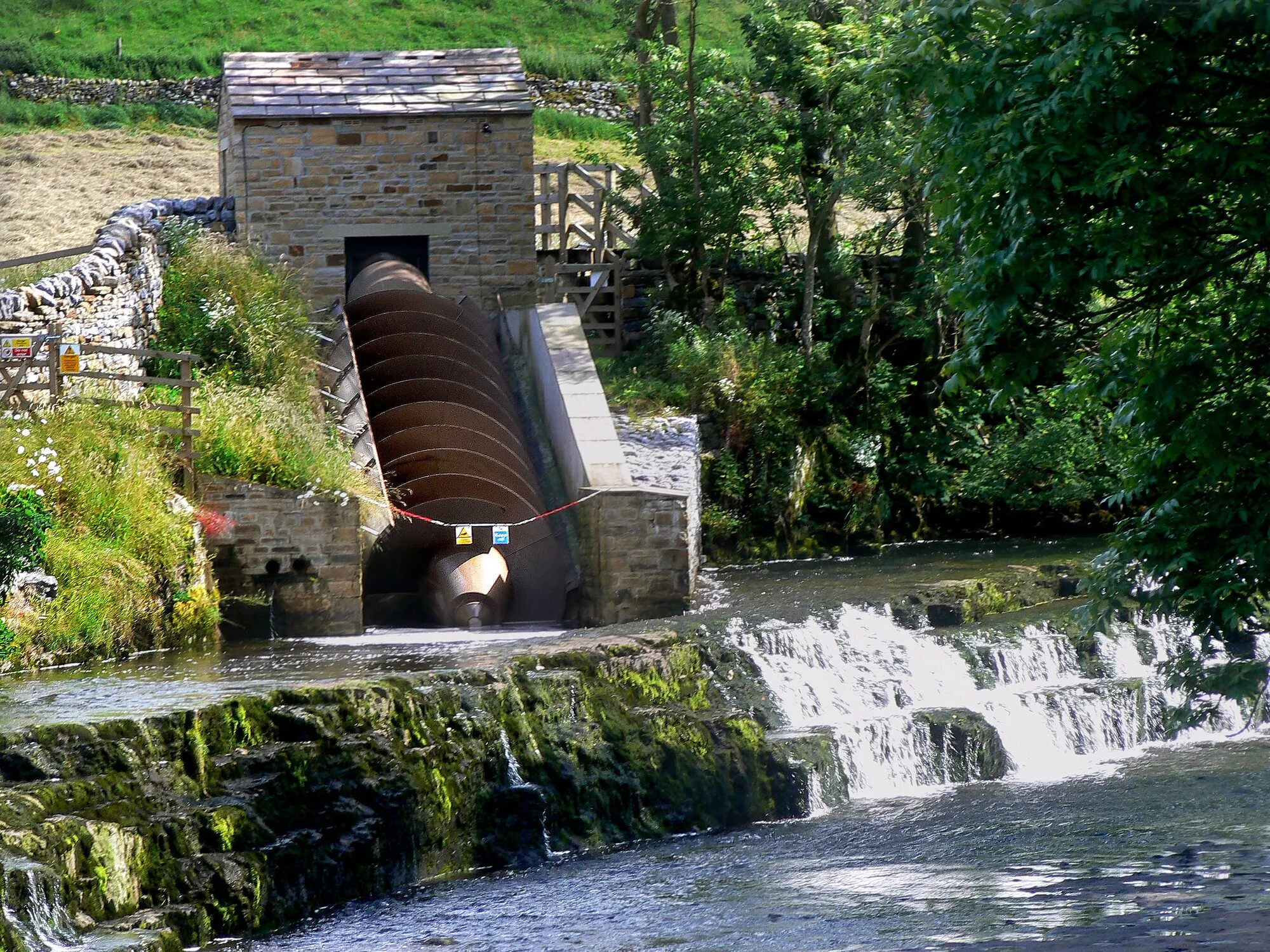 A large Archimedes screw in a stone channel beside a small waterfall, below a stone building among trees.
