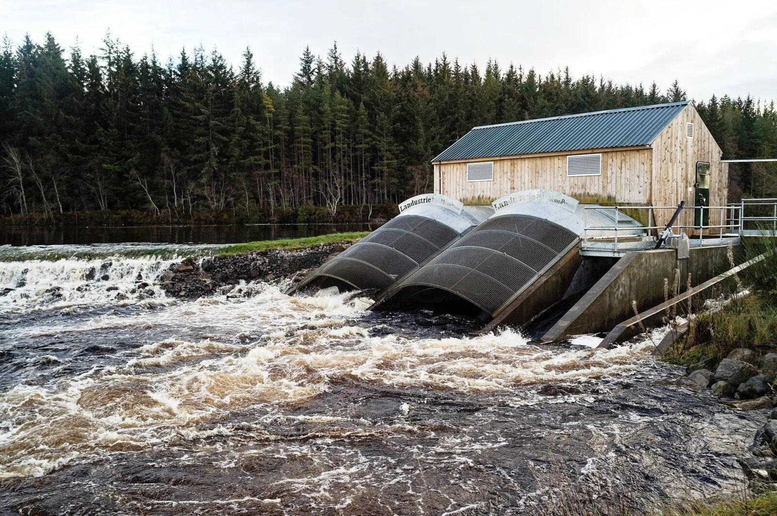 Two large Archimedes screws side by side at a river weir, with white water rushing out below them and a timber turbine house behind.