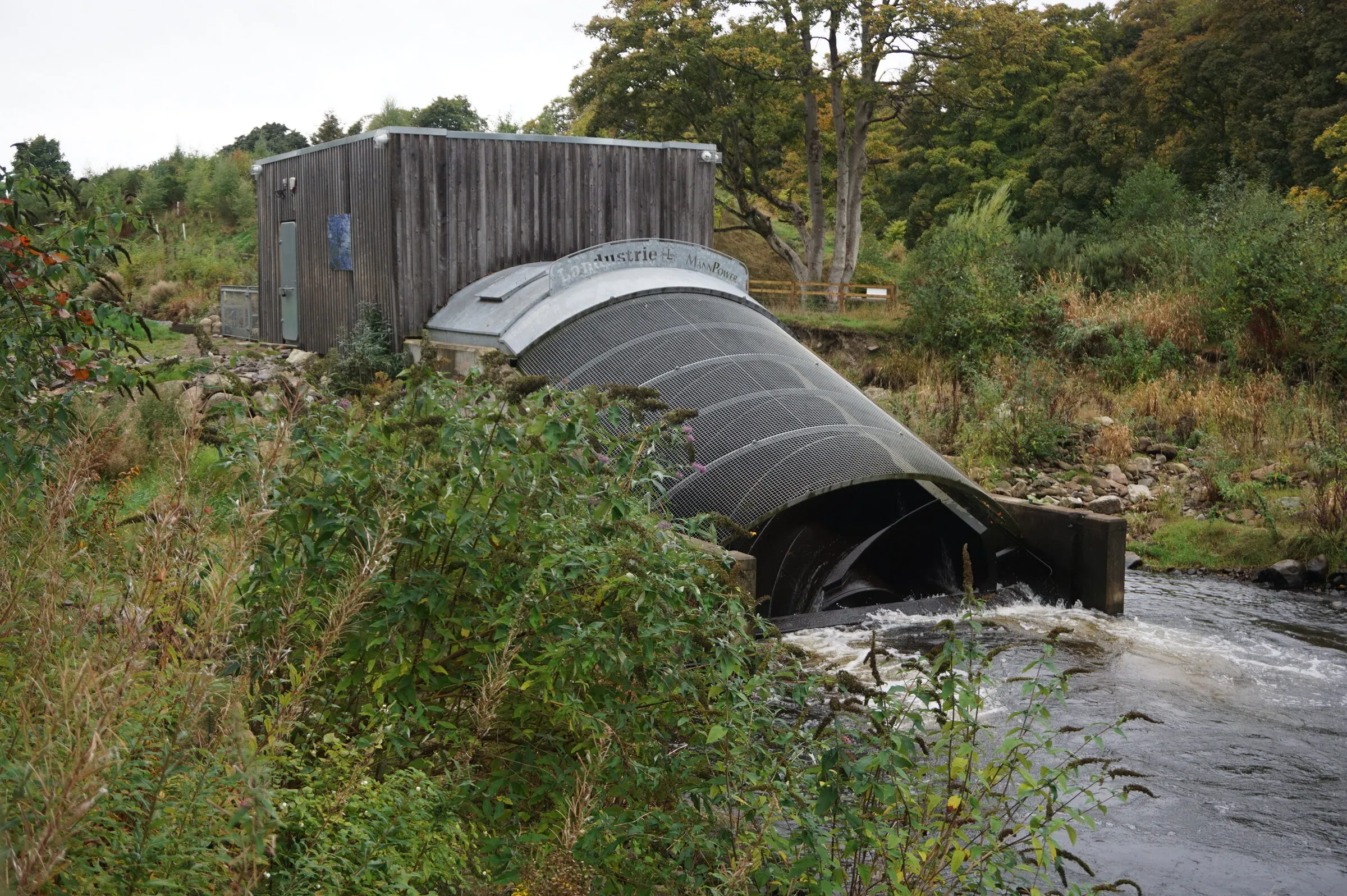 A large Archimedes screw turbine in a steel trough beside a river, with a timber turbine house behind it.