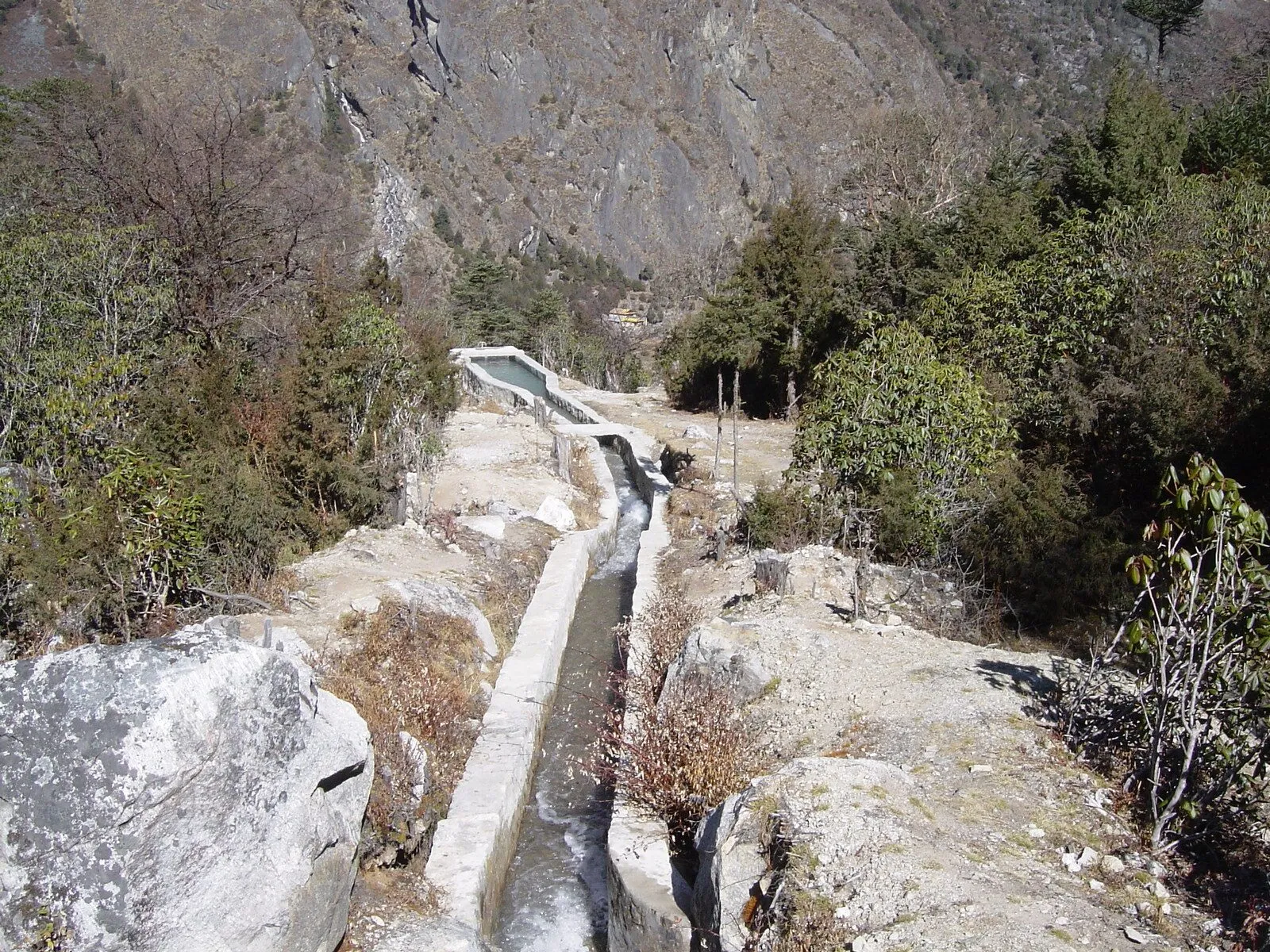 A narrow concrete canal running along a steep, rocky mountain valley towards a small basin.