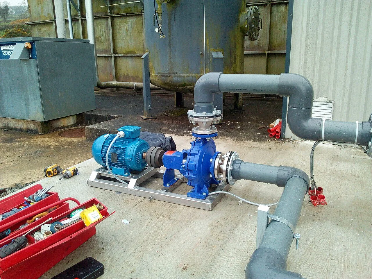 A blue pump coupled to a blue electric motor on a steel frame, connected to grey water pipes at a small water treatment site.