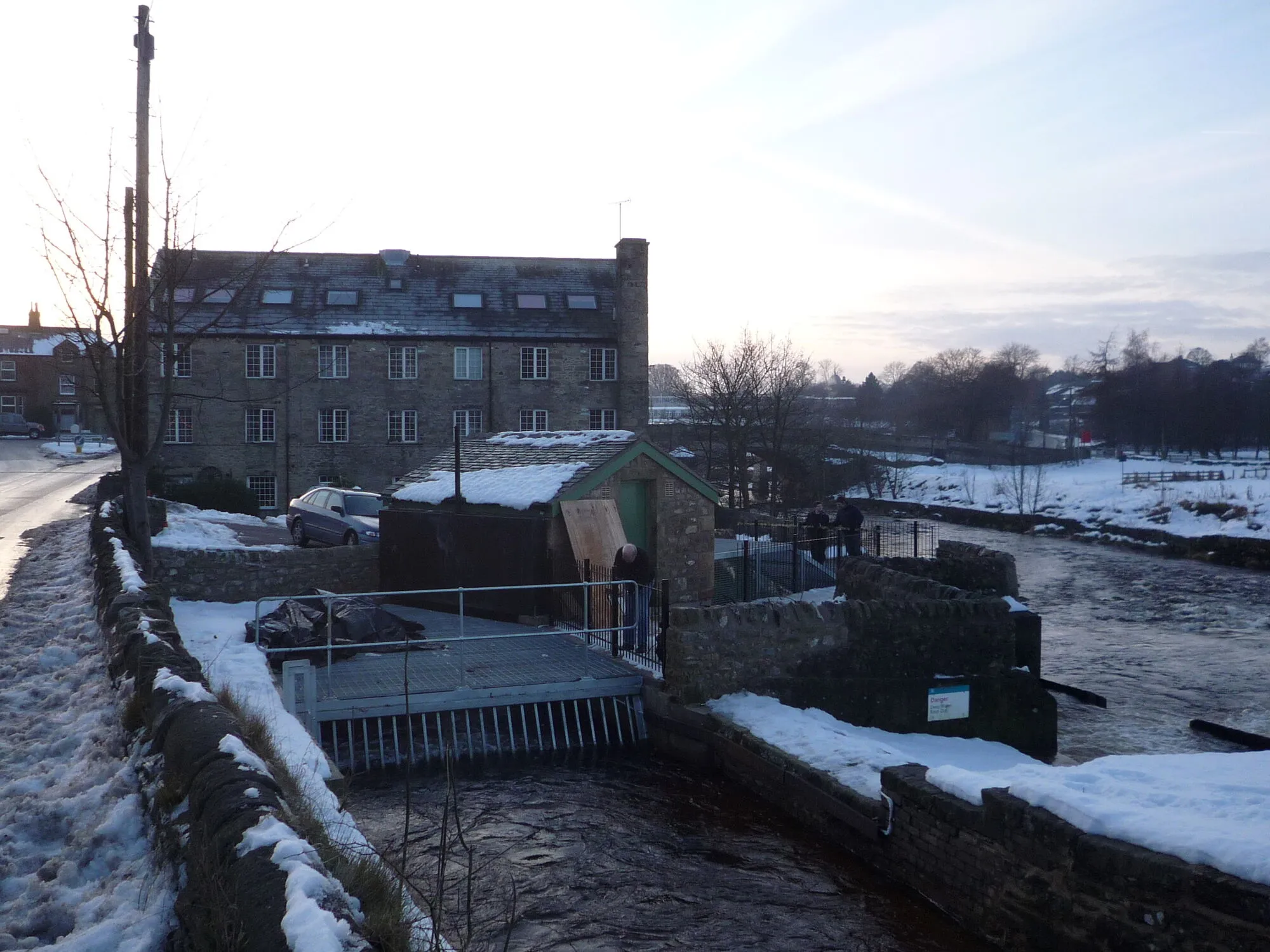 A small stone turbine house beside a snowy river weir in a Yorkshire town.