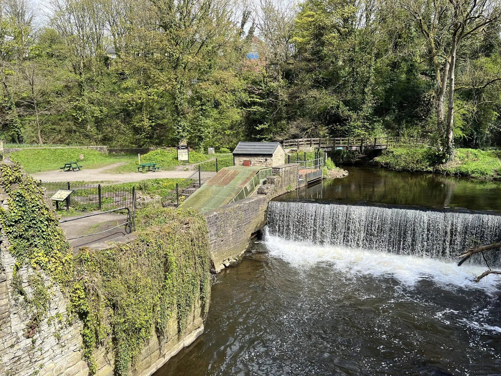 A curved stone weir across a river in a wooded gorge, with a small turbine house beside it.
