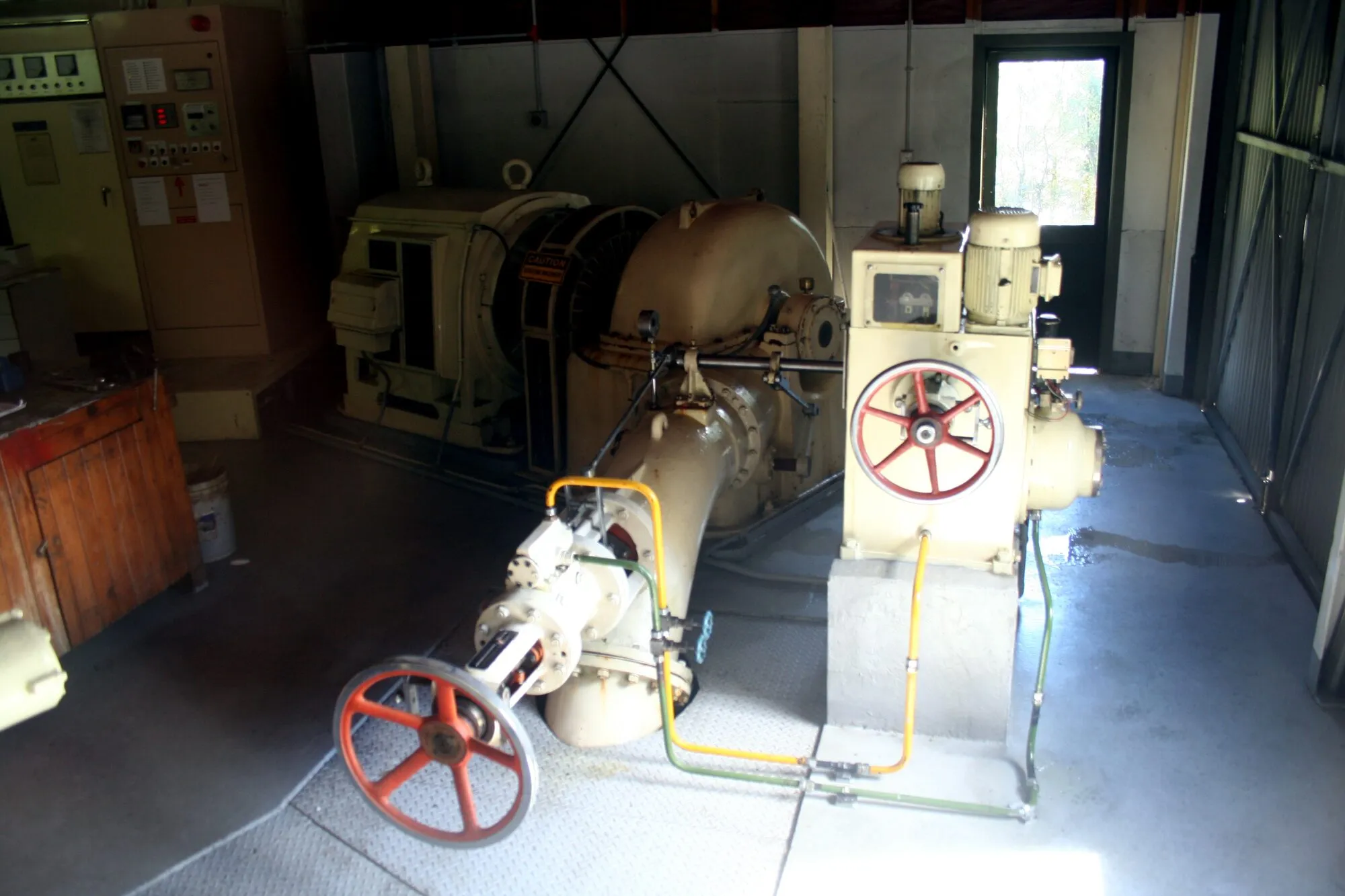 A small turbine and generator with red valve wheels and yellow pipework inside a compact powerhouse.