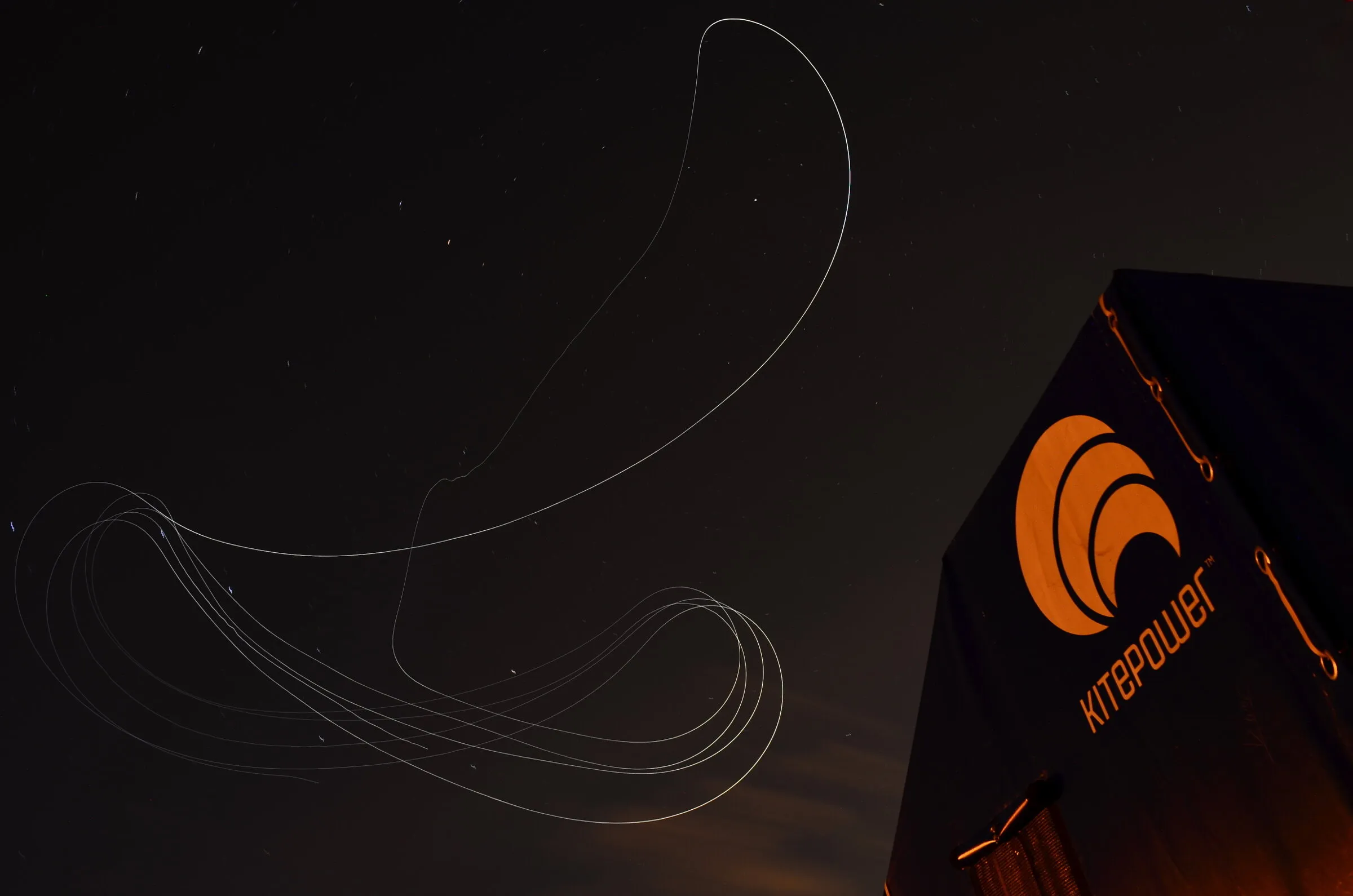 A long-exposure night photograph of a power kite's tracing light drawing figure-of-eight loops and a long return arc above a ground station.