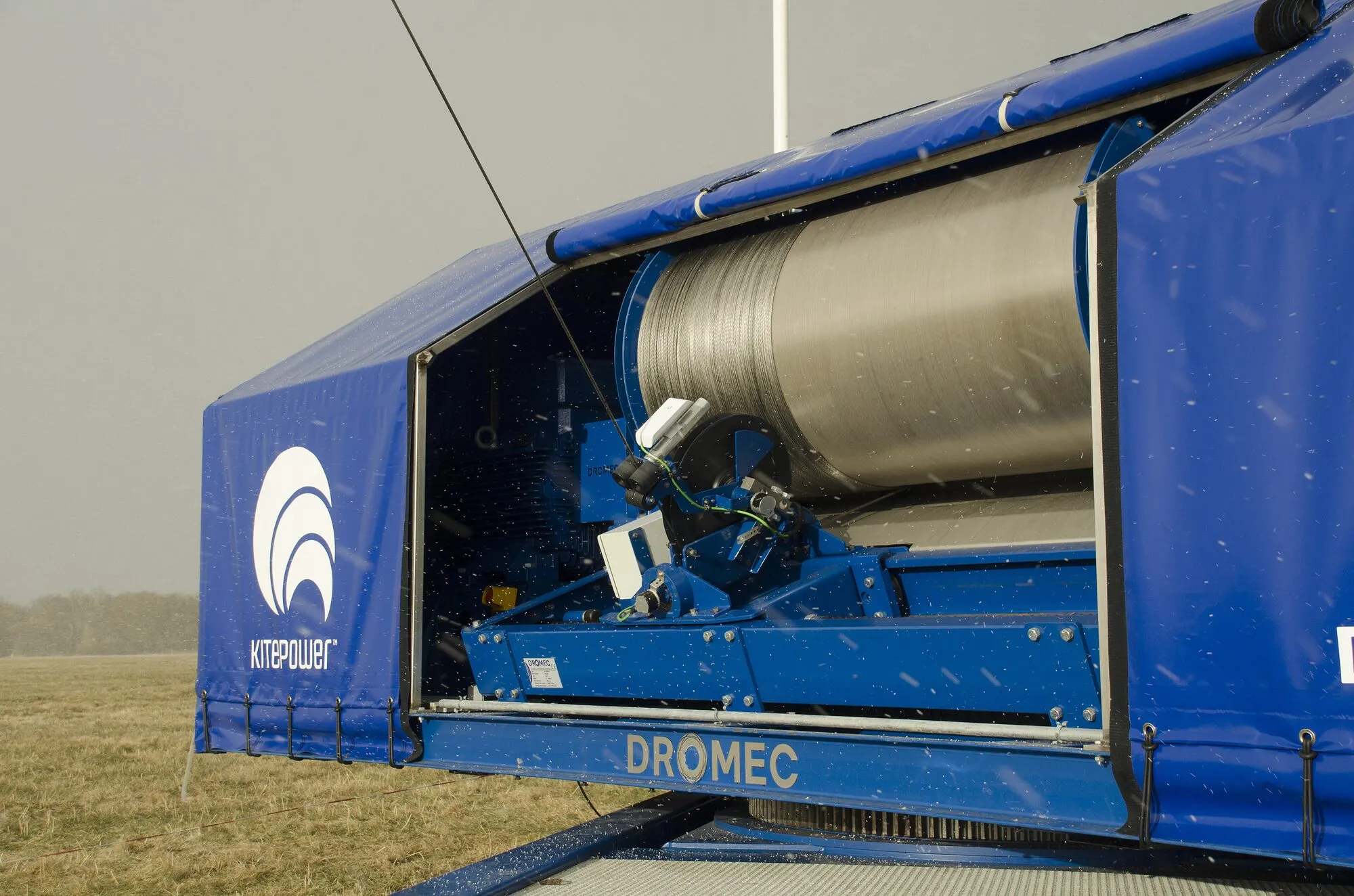 A blue container with its side opened, showing a large silver drum wound with tether, standing on a grassy field.