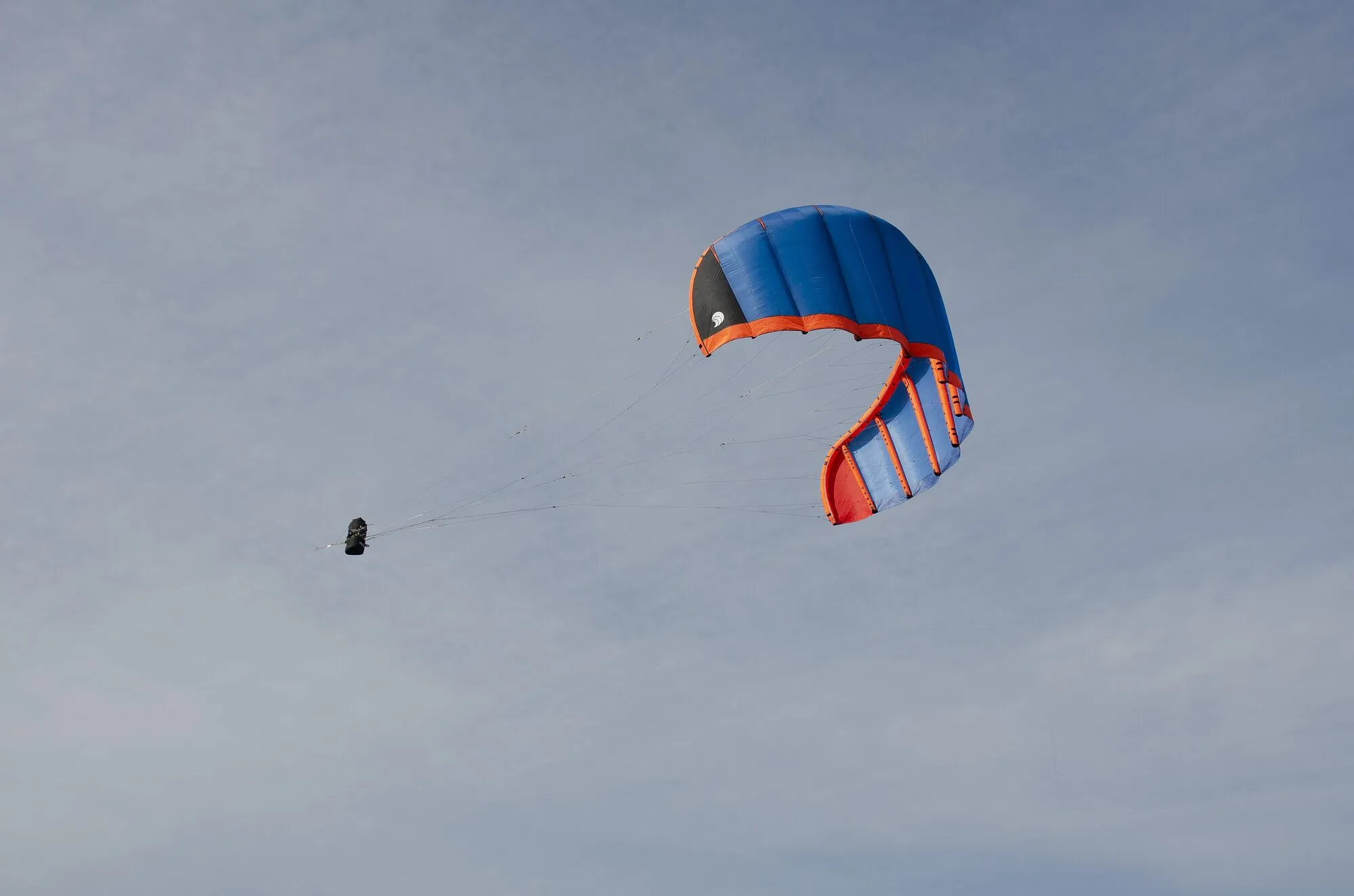 A red and blue soft kite high in a grey sky, with a small control box hanging on its lines below the wing.