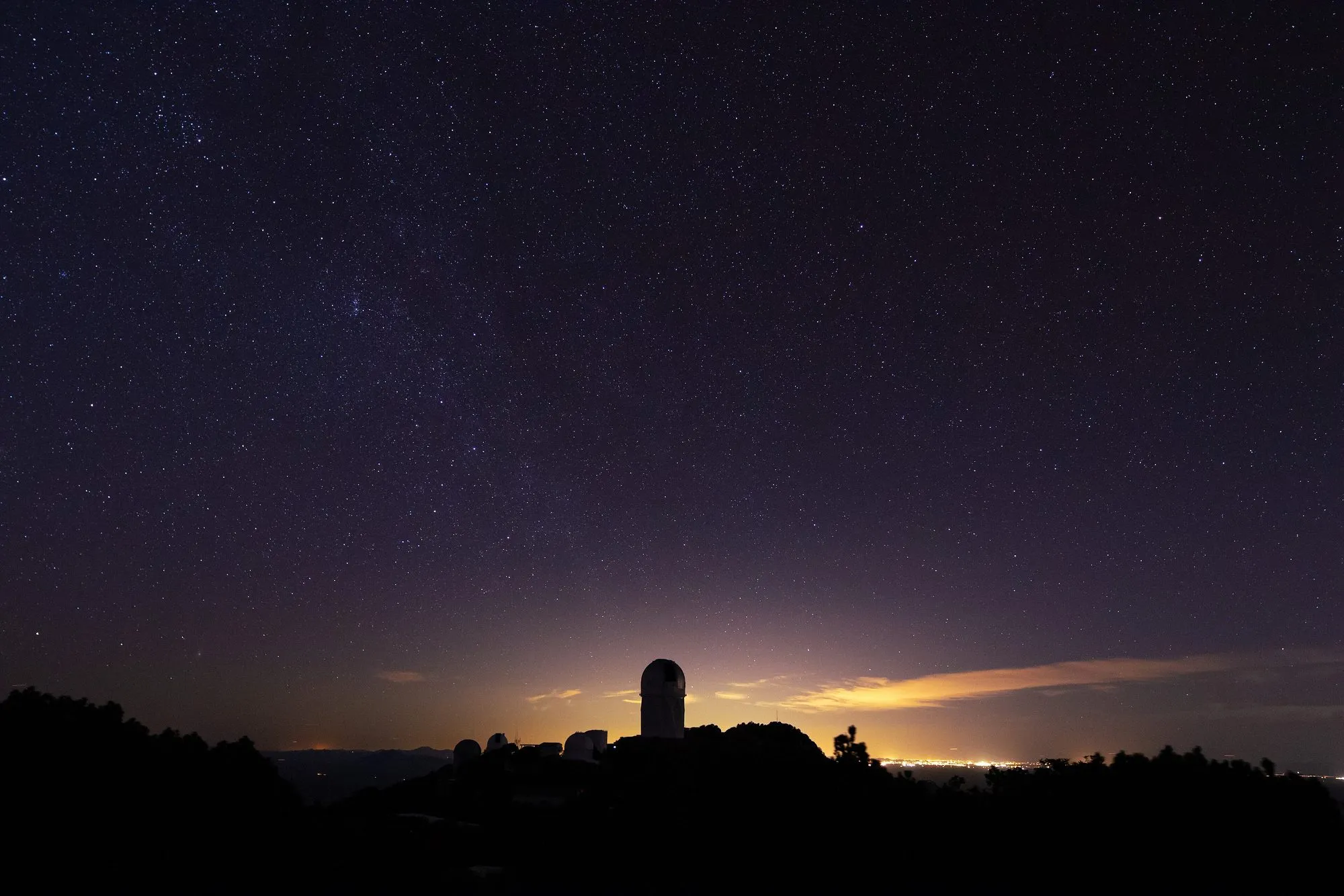 Die gloed van 'n stad se ligte op die horison agter die koepels van Kitt Peak Nasionale Observatorium onder 'n sterrehemel.