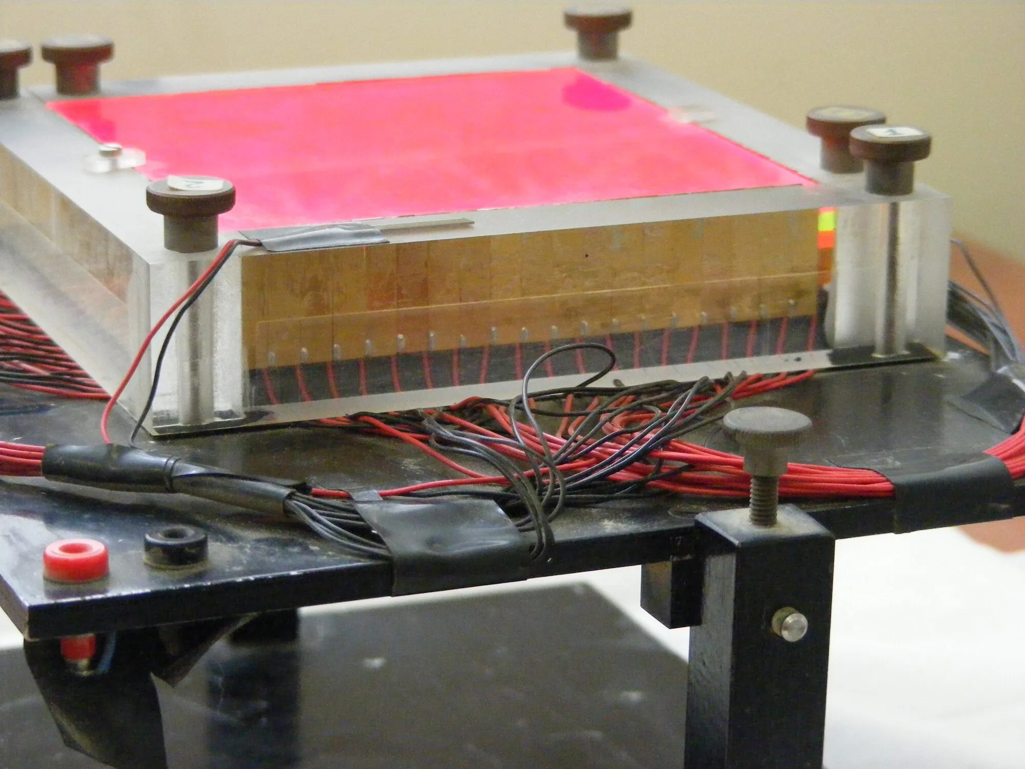 A square sheet of glowing pink plastic in a metal frame, with rows of small solar cells along its edges and wires leading away.