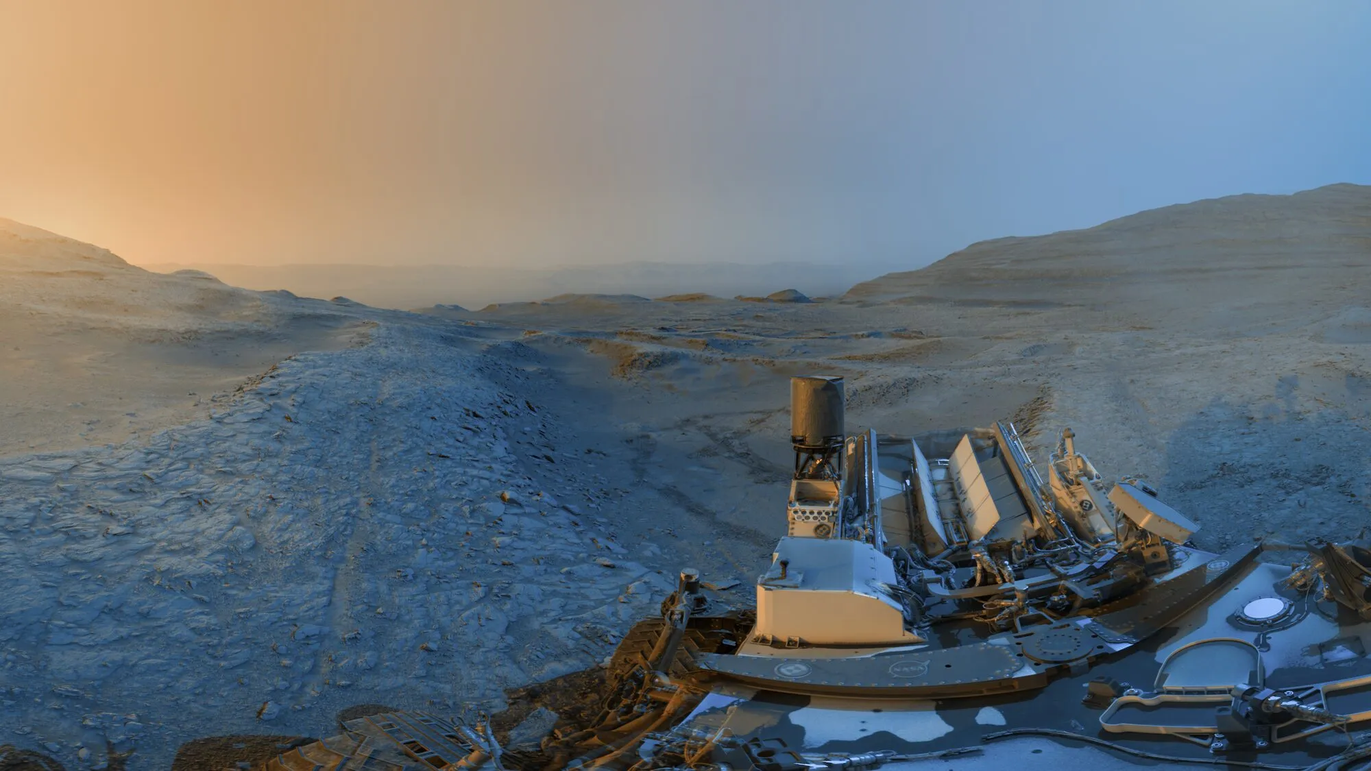 Una cresta rocosa marciana sota un cel que passa de taronja a blau, amb la plataforma del rover Curiosity en primer pla i turons a la distància.