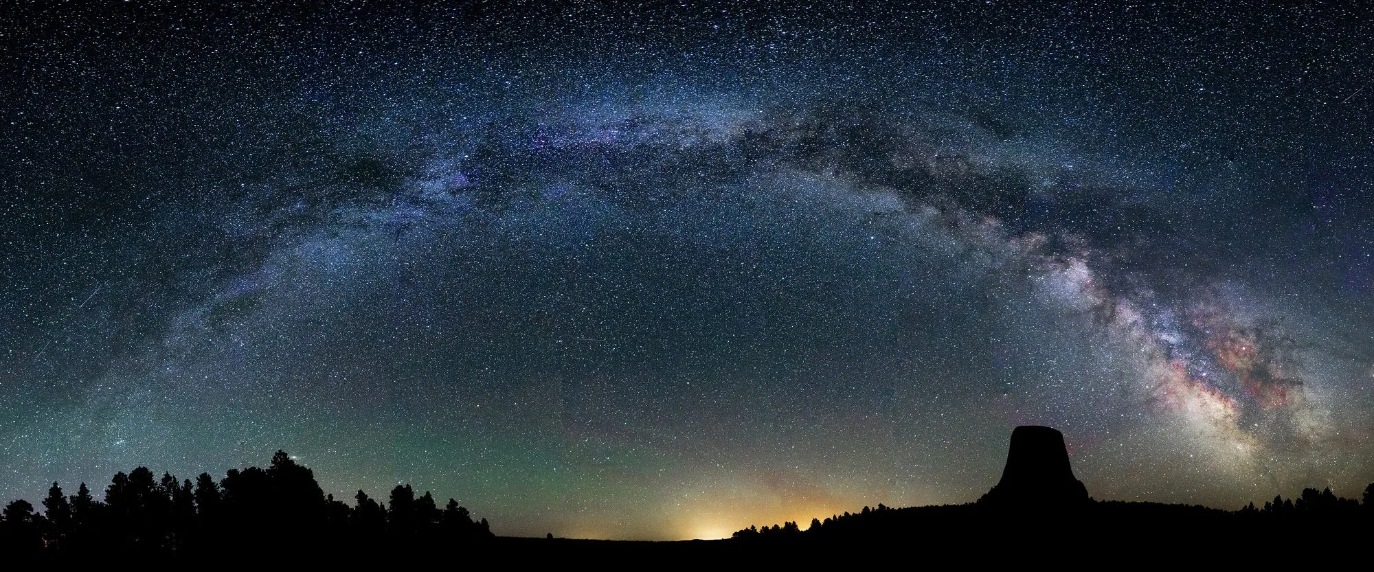 La Via Làctia en arc a través d'un cel estrellat sobre un bosc fosc i la silueta de Devils Tower, Wyoming.