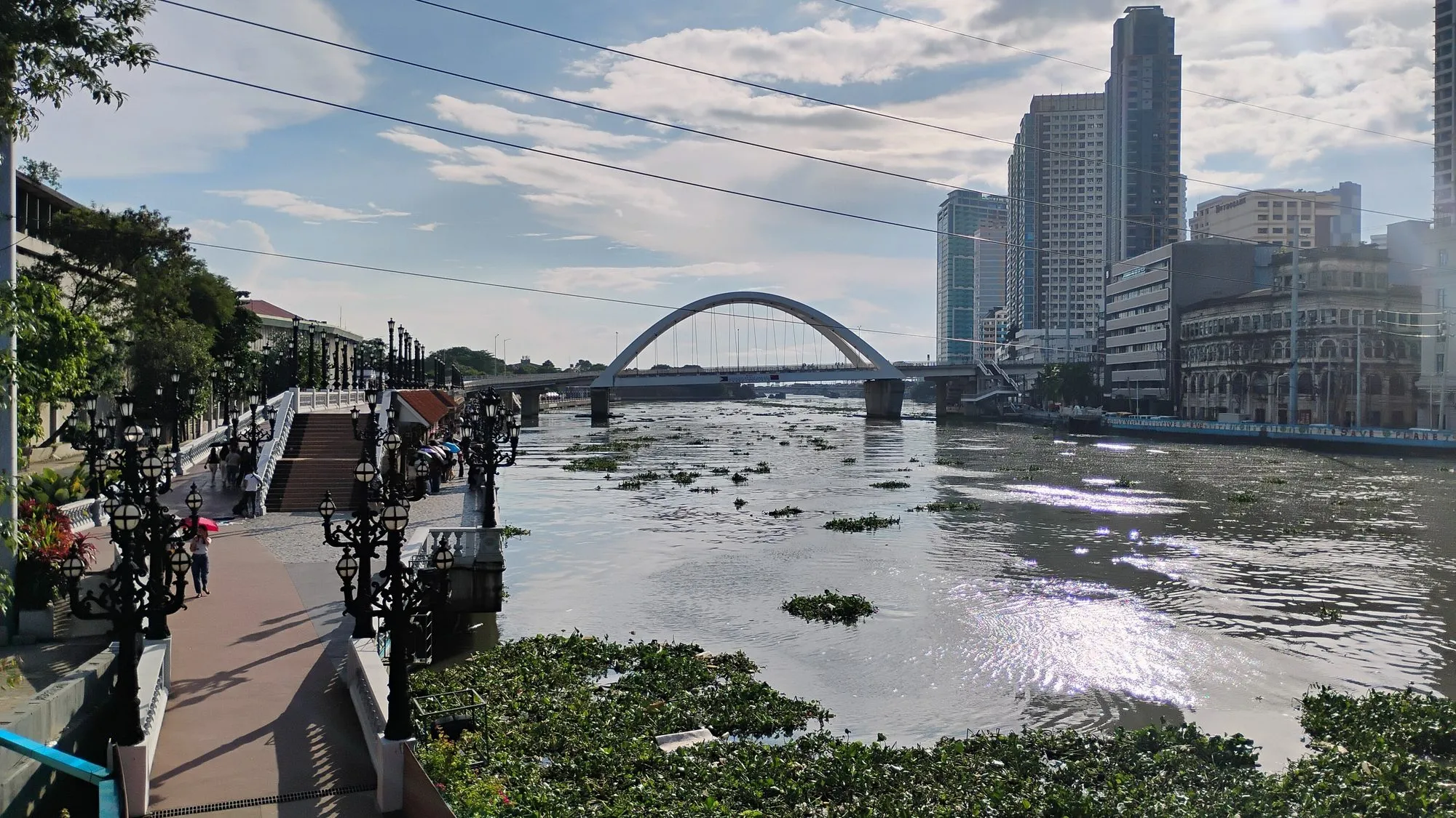 Un passeig fluvial amb lluminàries ornamentades al costat del riu Pasig a Manila, amb jacints d'aigua sobre l'aigua i un pont més enllà.