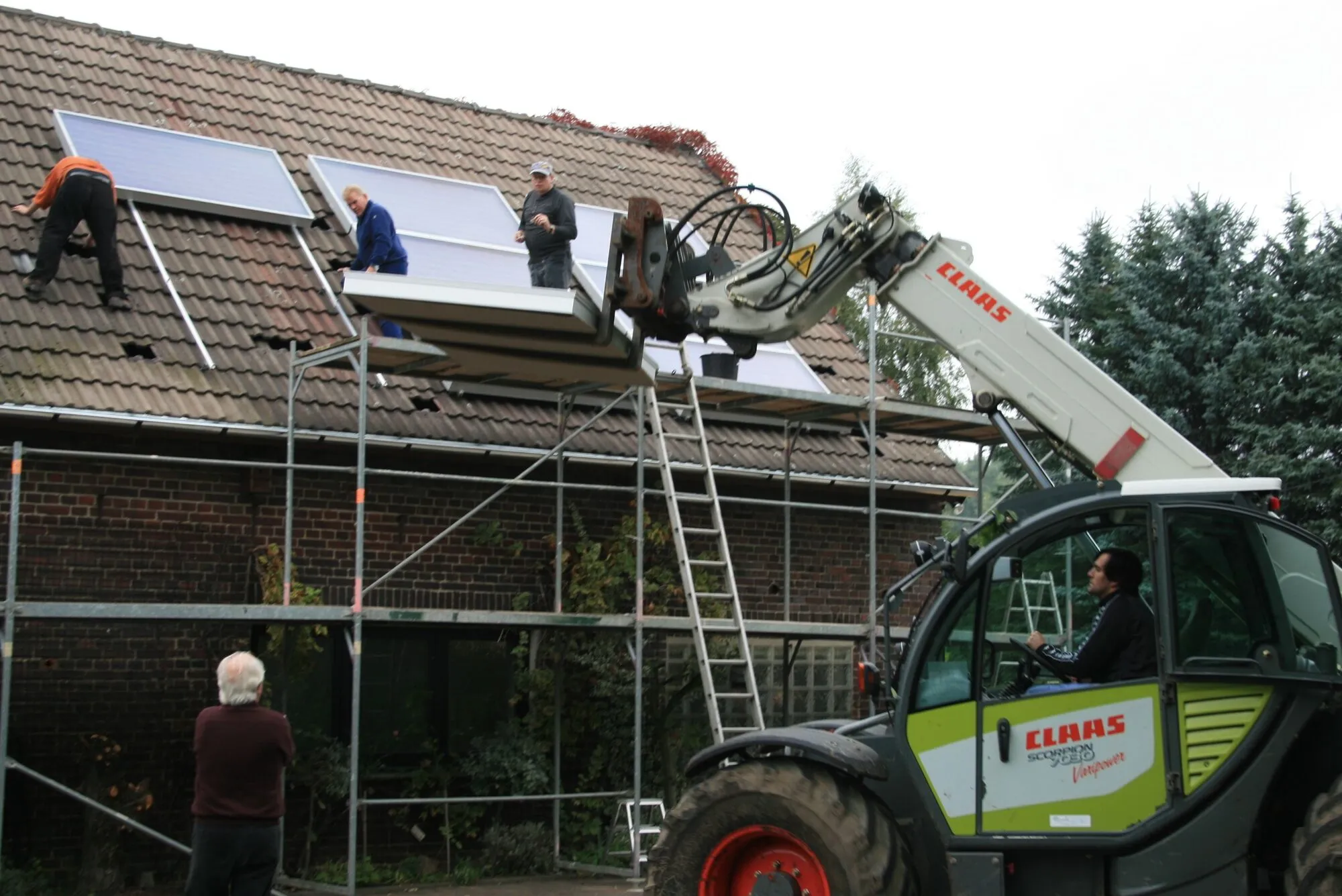 Workers on scaffolding lift a large flat solar collector onto a tiled roof with a telehandler while others guide it from below.