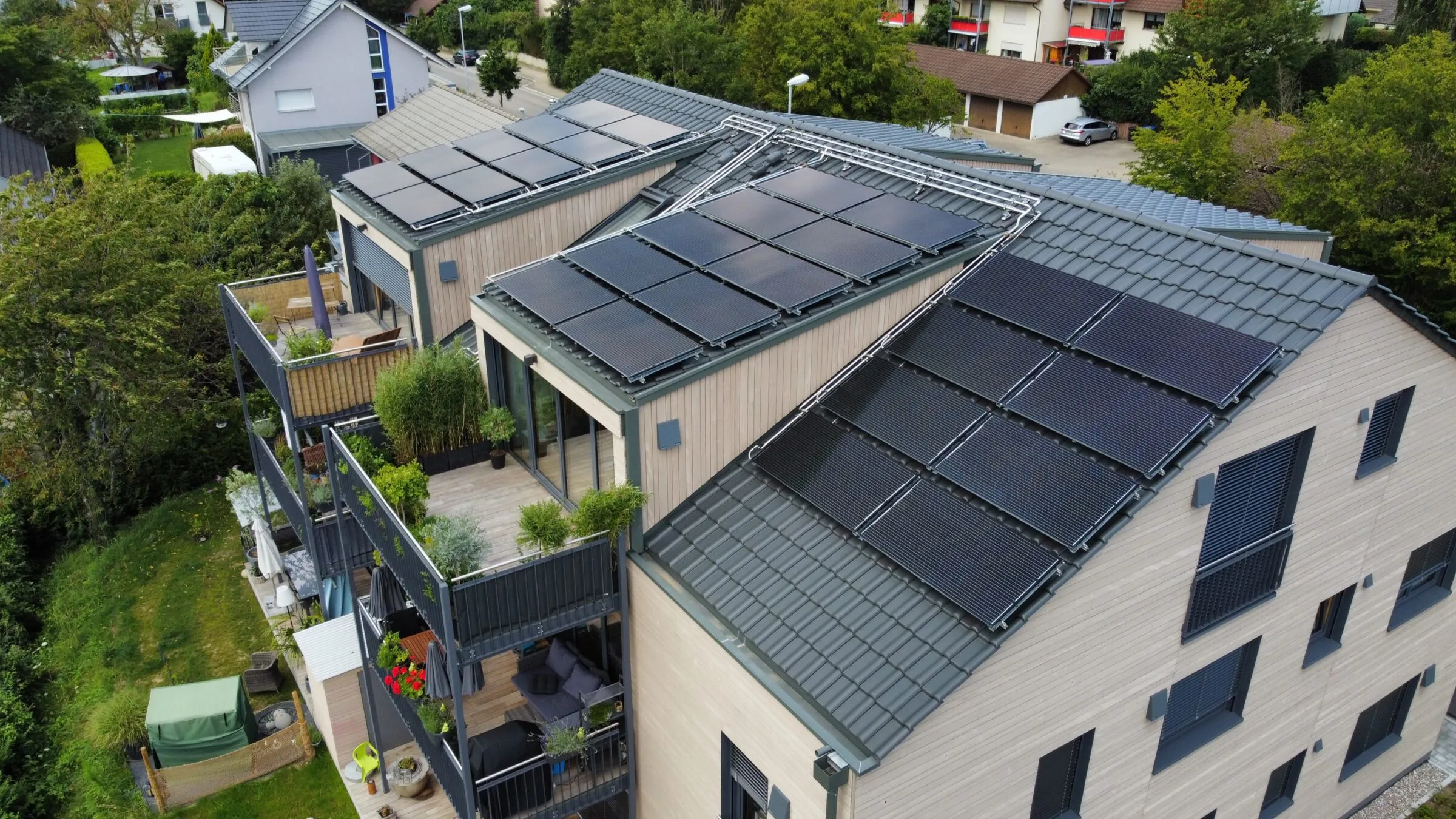 Dark PV-thermal collectors covering the pitched roofs of a modern apartment building among gardens.