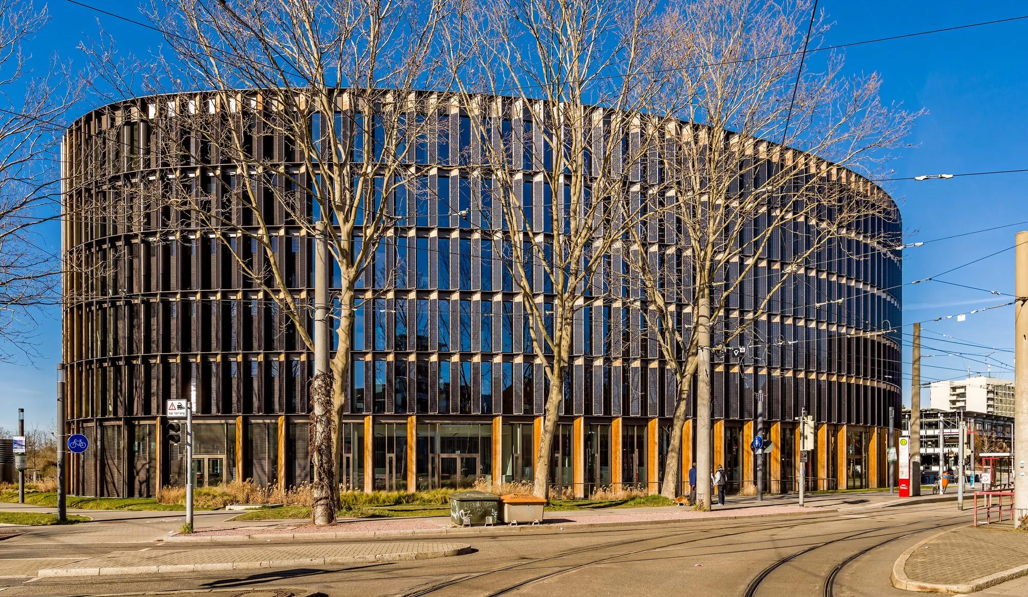 A round, seven-storey office building clad in vertical timber slats and dark glass, with bare trees and tram tracks in front.