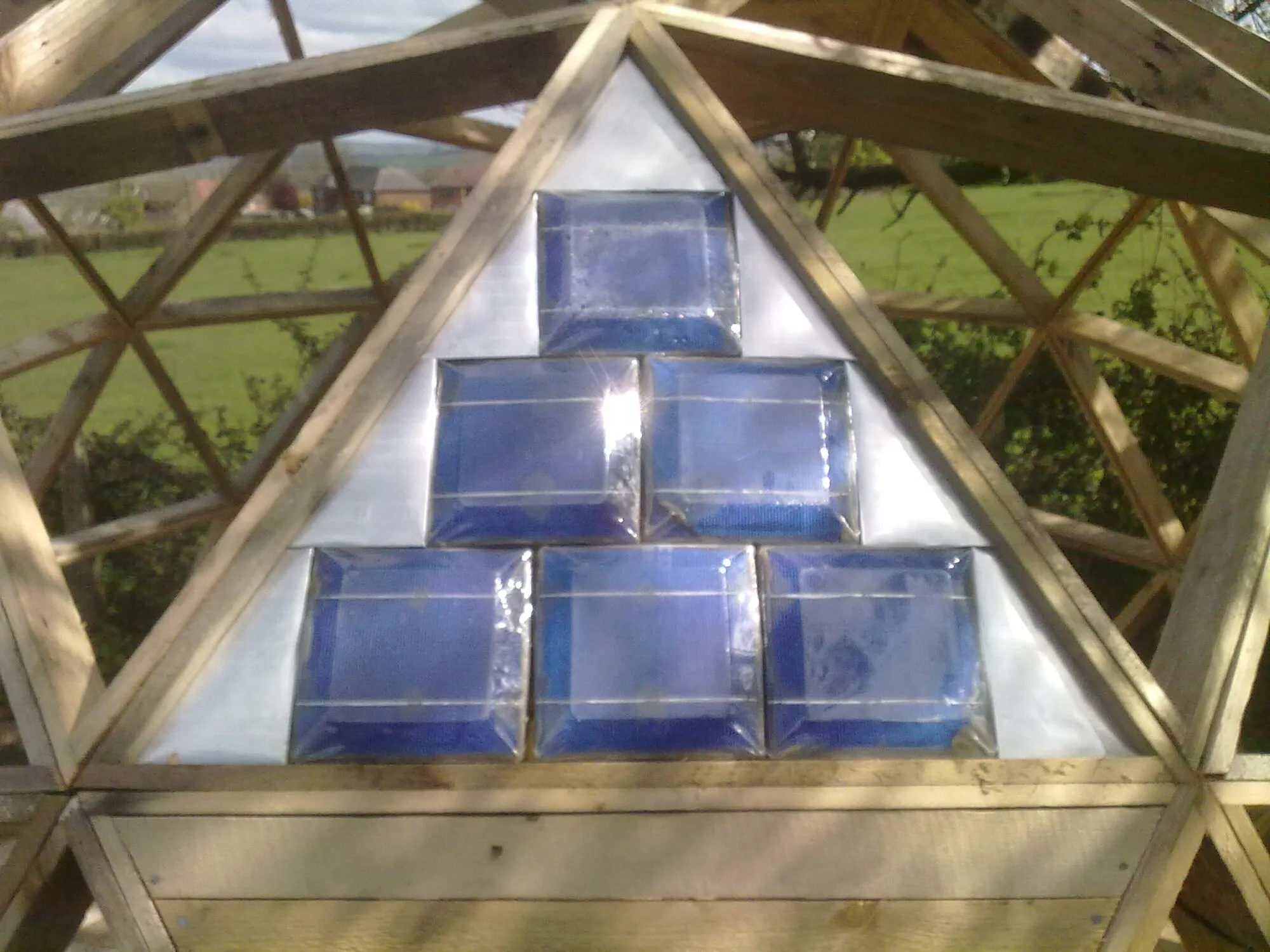 Small solar cells mounted on a triangular metal absorber inside the timber frame of a geodesic dome, with a field behind.