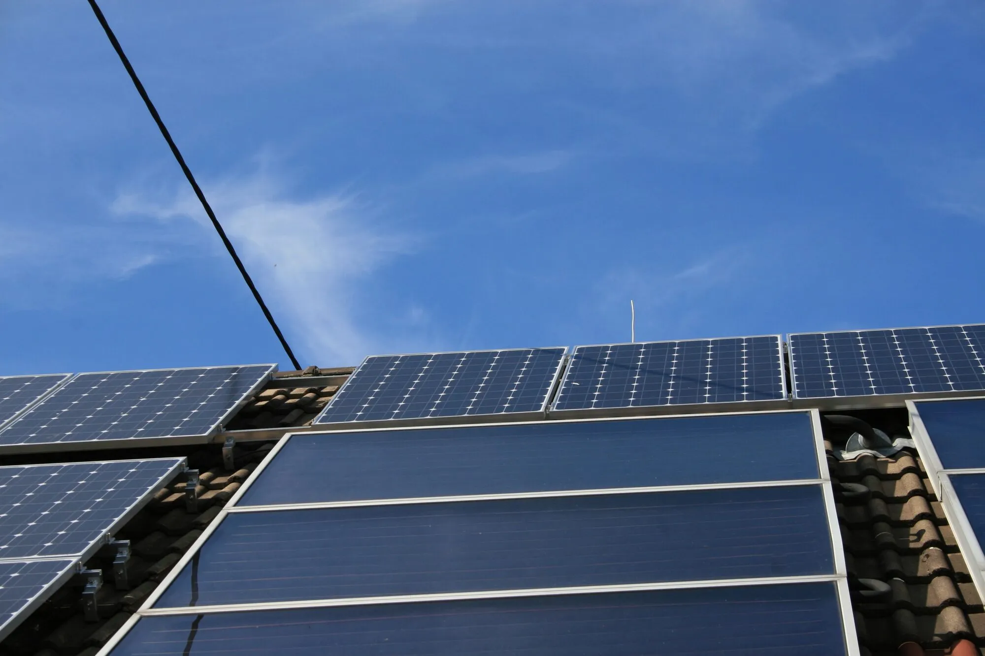 A tiled barn roof carrying dark solar panels at the top and a row of flat solar thermal collectors below them, against a blue sky.