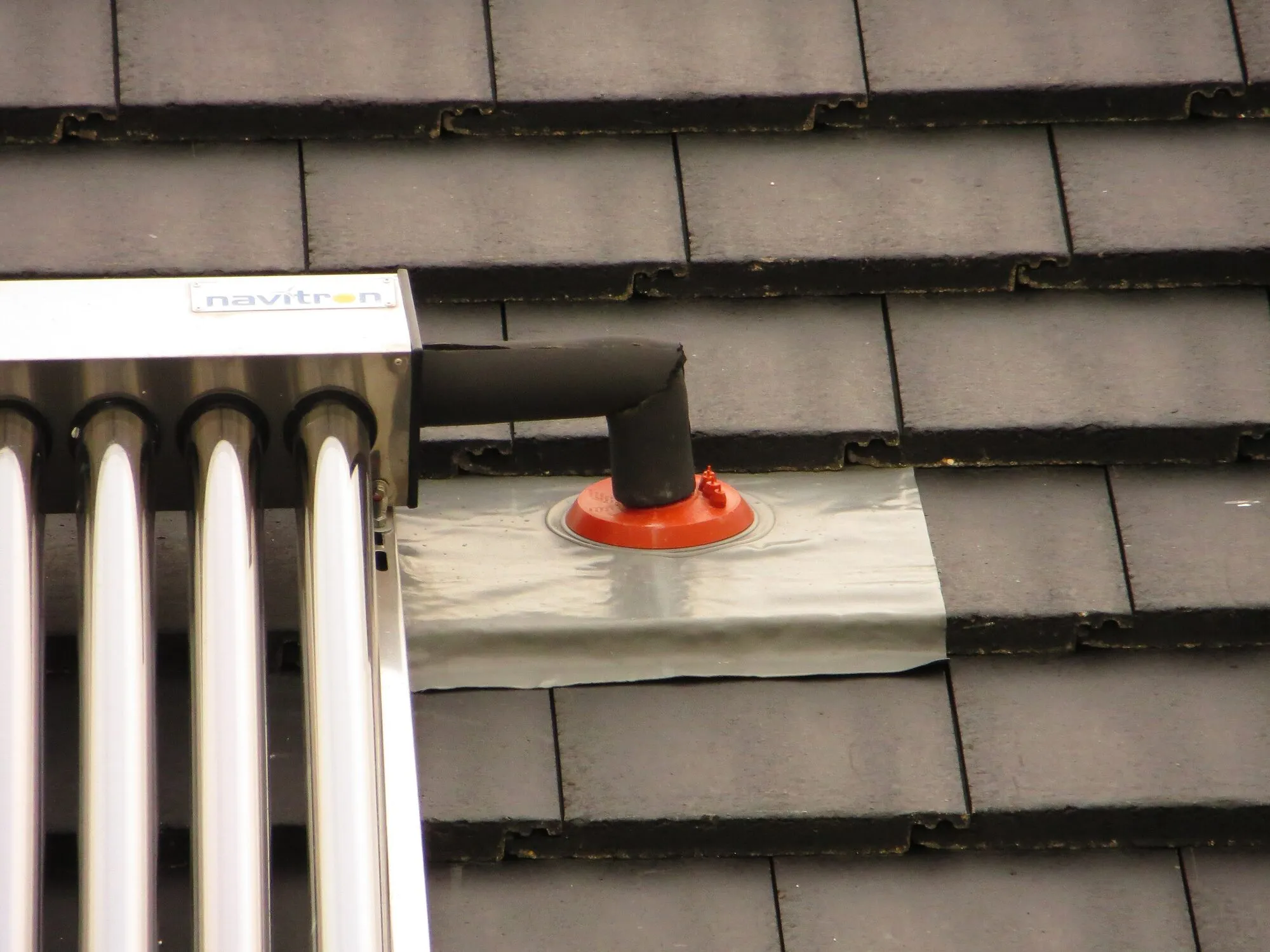 Close-up of insulated pipes rising from the end of a glass-tube solar collector and passing through a sealed flashing in a tiled roof.