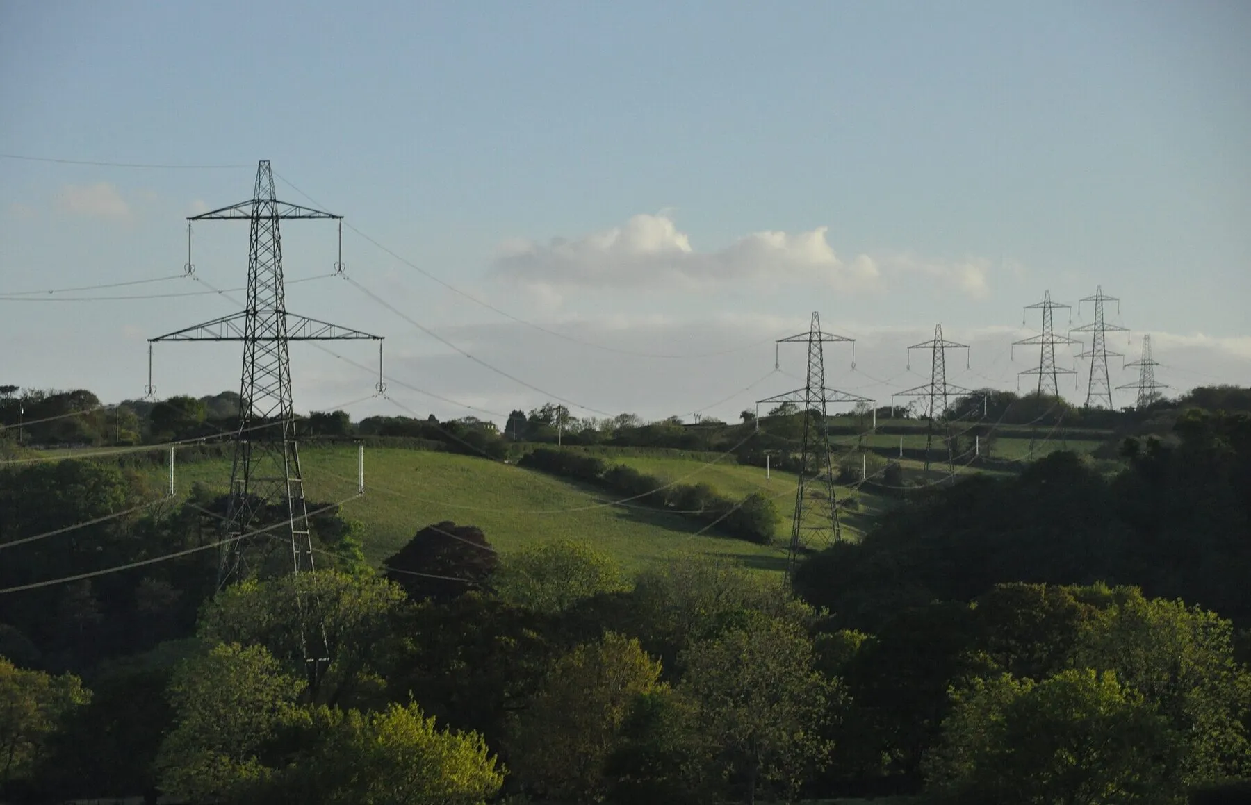 A high-voltage pylons crossing the green farmland and forests in Devon, England.