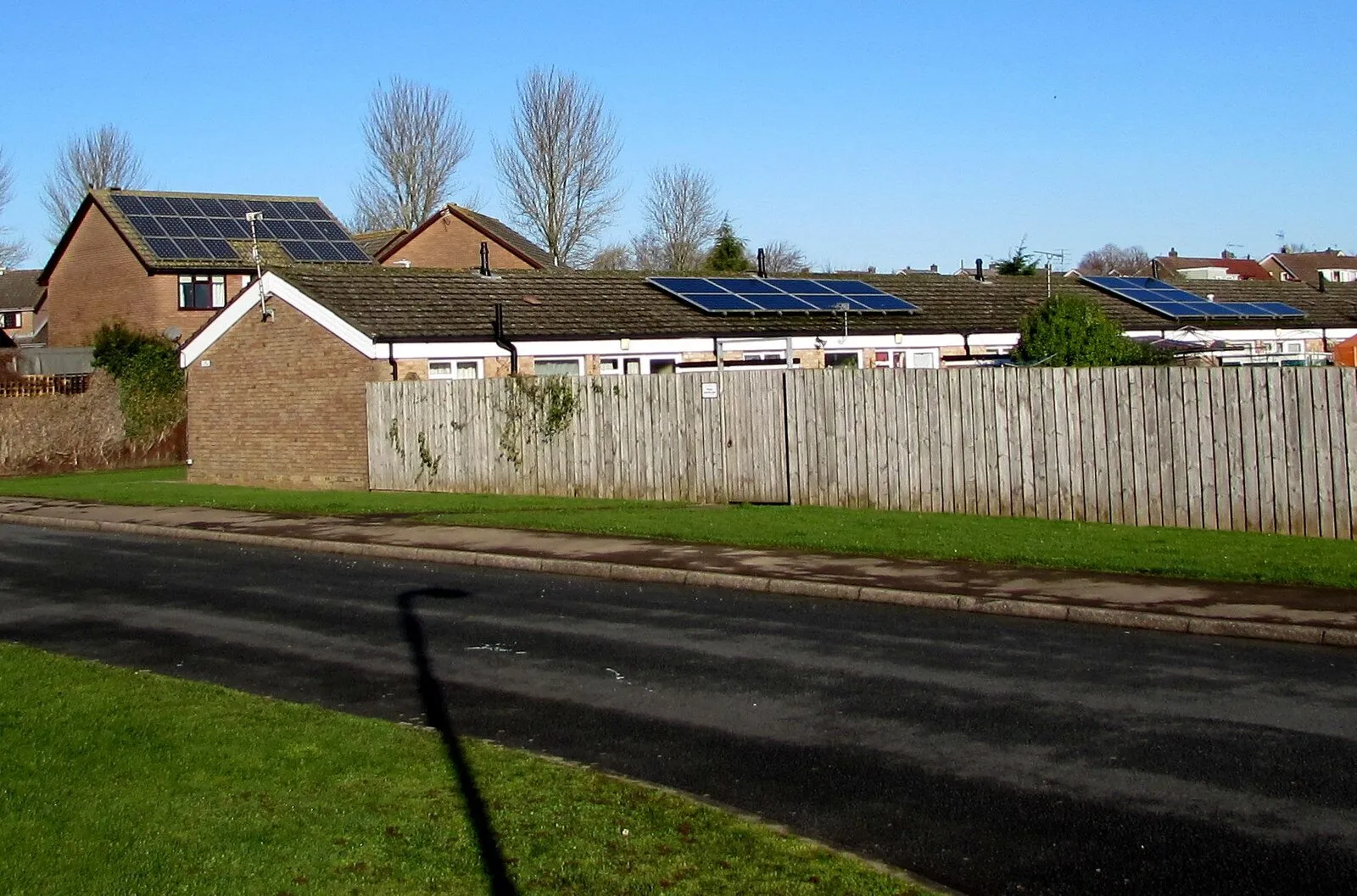 Solar panels on the roofs of a row of single-storey houses beside a road and a green verge.